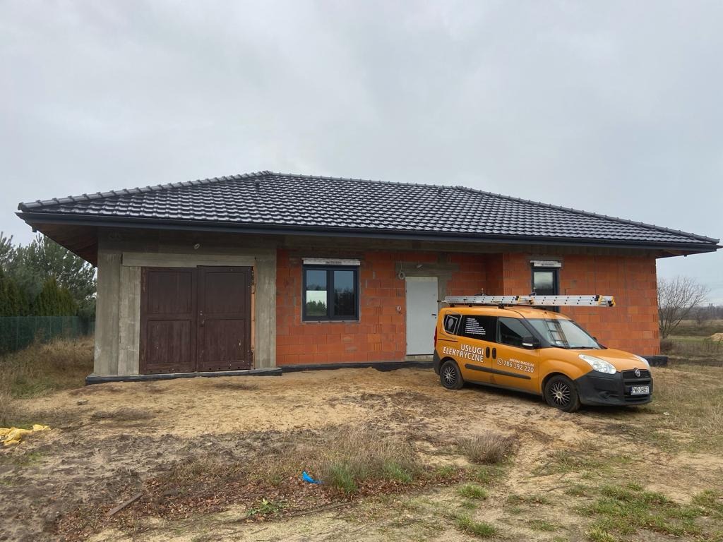 Pomarańczowy samochód z napisem 'Usługi Elektryczne' zaparkowany przed surowym budynkiem z cegły z czarnym dachem, drabina na dachu pojazdu.