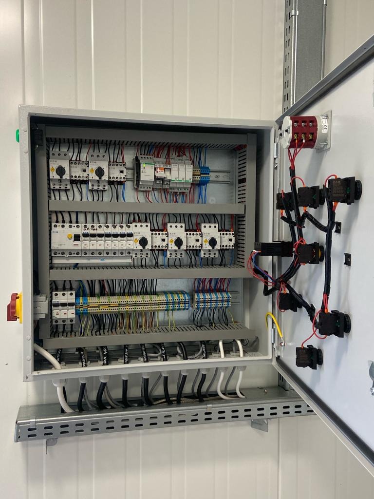 Otwarta szafa sterownicza z widocznymi elementami elektrycznymi, takimi jak przekaźniki, bezpieczniki i okablowanie, z żółto-zielonym przewodem ochronnym po prawej stronie.