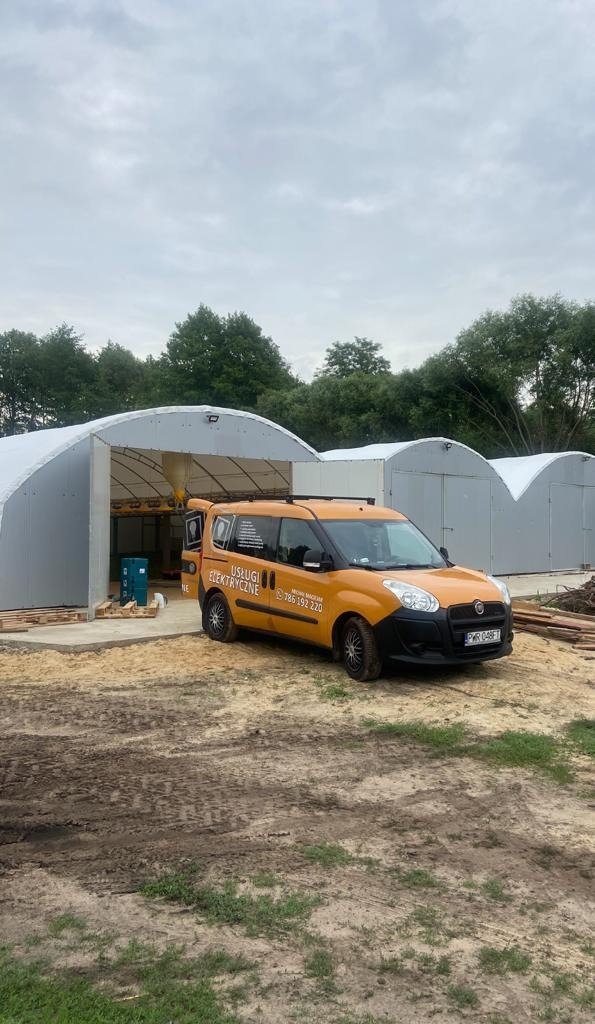 Pomarańczowy samochód z napisem 'Usługi Elektryczne' zaparkowany przed metalowymi halami łukowymi, z otwartymi drzwiami po stronie kierowcy.