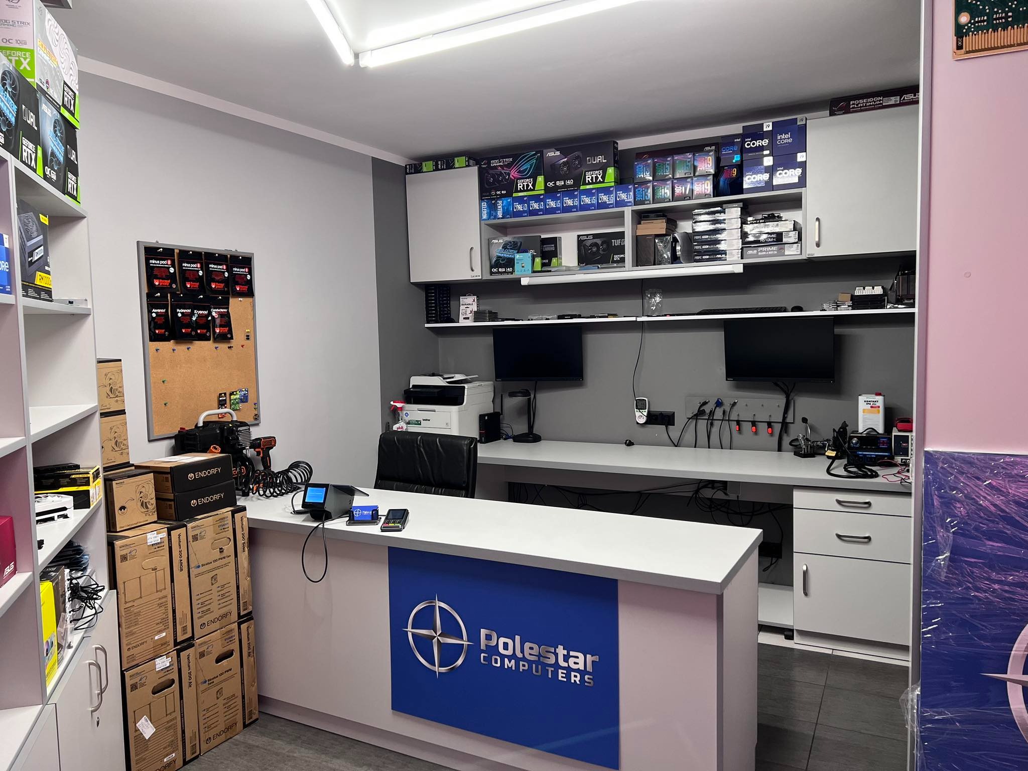 Wnętrze serwisu komputerowego z biurkiem recepcyjnym oznaczonym logo Polestar Computers, półki z komponentami komputerowymi, narzędzia na blacie i sprzęt diagnostyczny.
