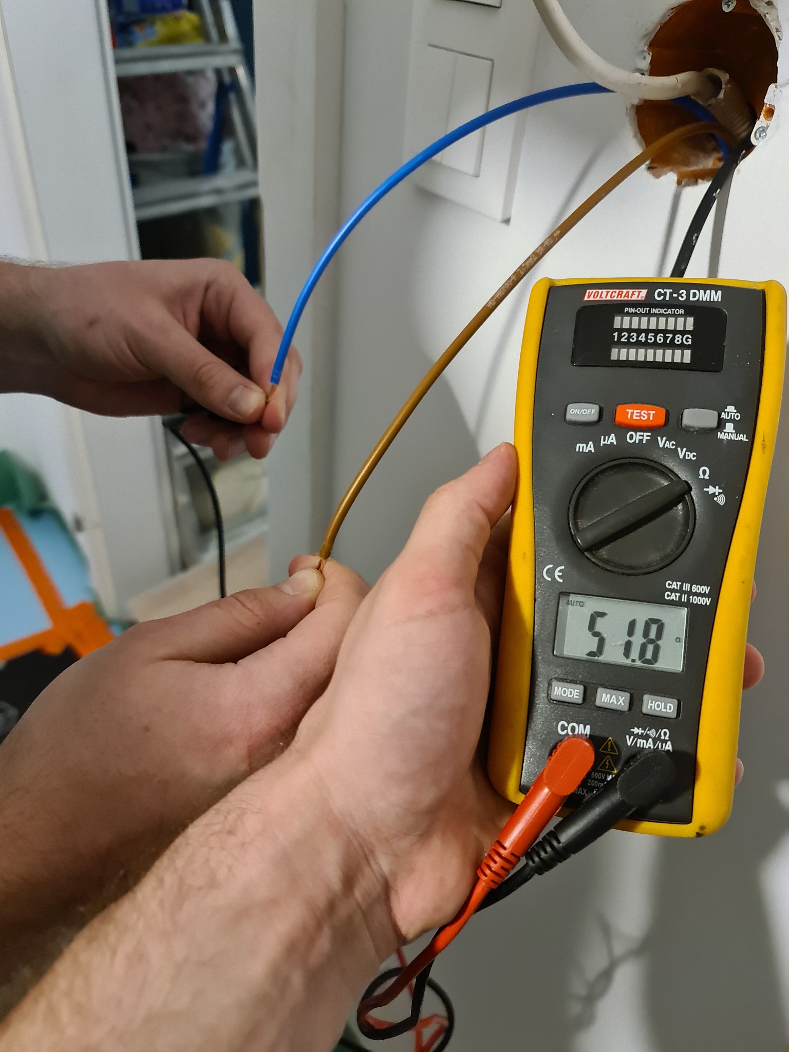 Pomiar napięcia w instalacji elektrycznej za pomocą multimetru Volcraft CT-3 DMM, widoczne przewody: niebieski i brązowy, obok otwór w ścianie z wystającymi kablami.