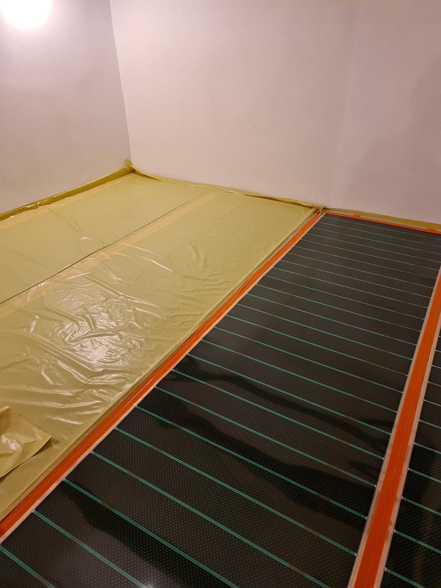 Fragment podłogi z częściowo rozłożoną matą grzewczą z zielonymi liniami, obok folia ochronna na podłożu, widoczne listwy przypodłogowe.