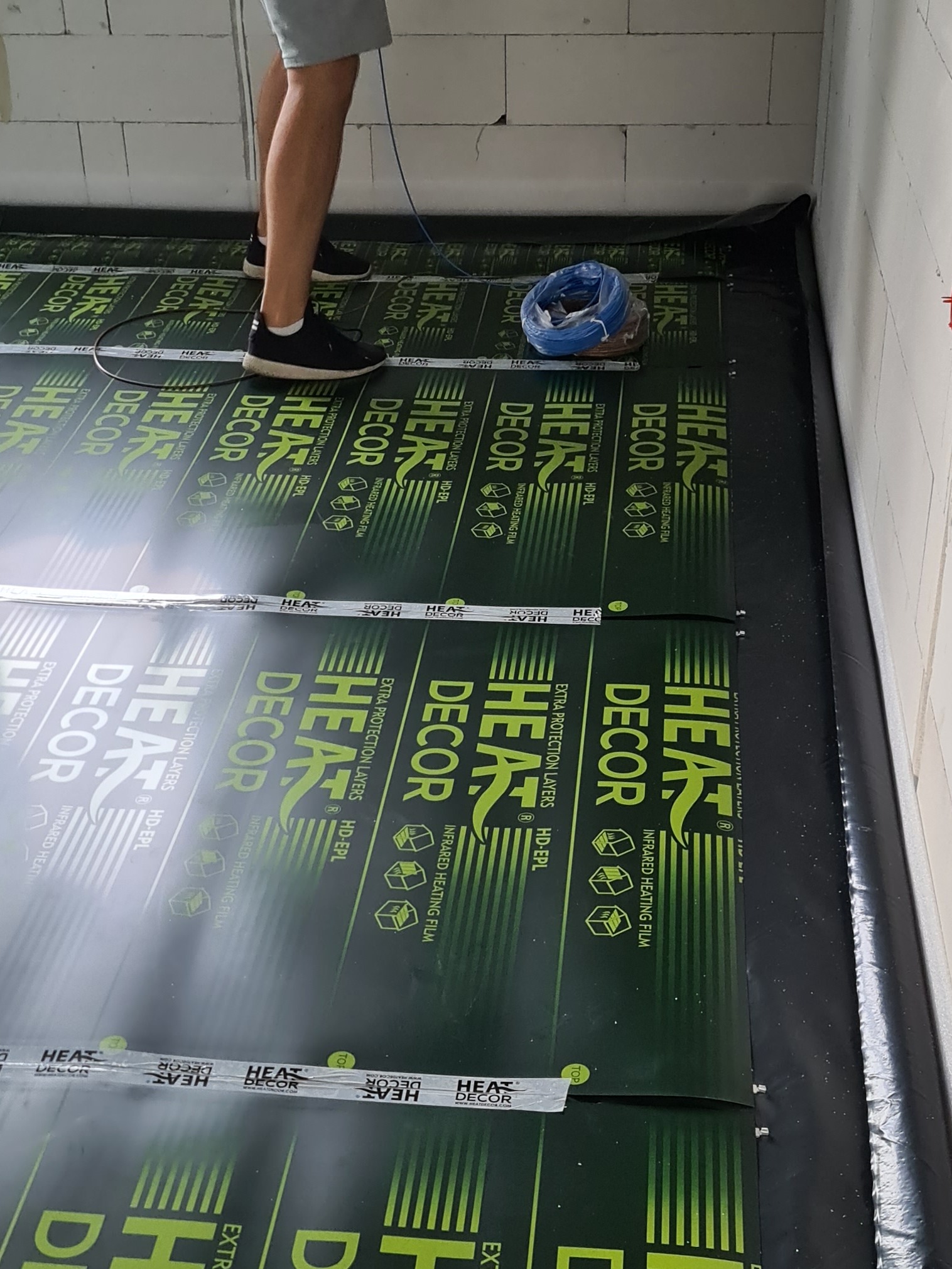 Montaż folii grzewczej pod podłogę w Stalowej Woli, widoczne rozłożone arkusze z nadrukiem 'HEAT DECOR' oraz zwój niebieskiego przewodu elektrycznego.