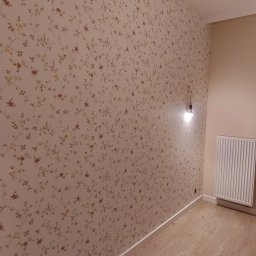 Jacek Brzezicki - Ściana z tapetą w drobne kwiaty, widoczna instalacja elektryczna z żarówką i grzejnik na ścianie w wykończonym pokoju.