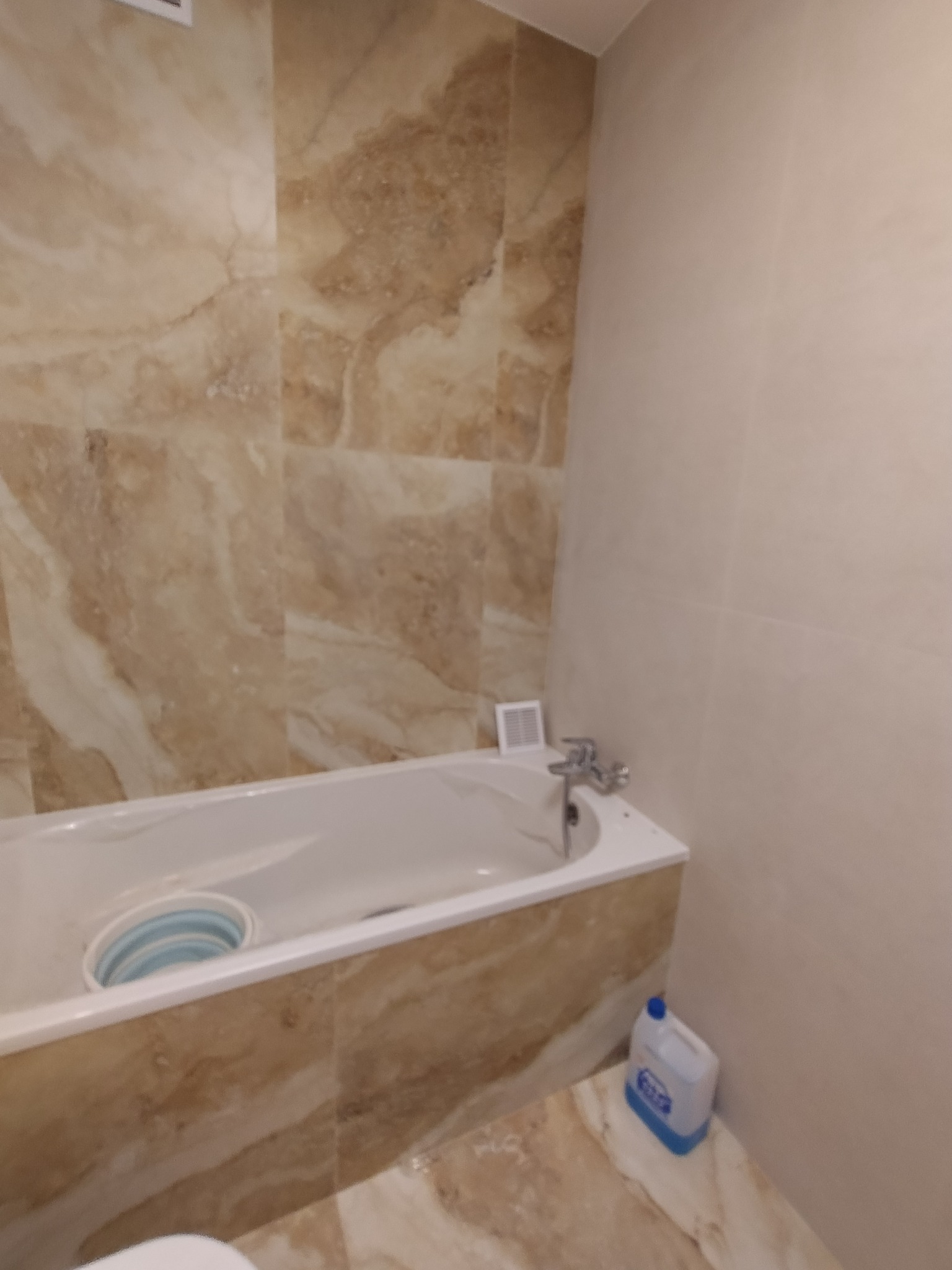 Wykończona łazienka z wanną obłożoną płytkami imitującymi marmur, widoczna składana miska w wannie oraz butelka płynu do czyszczenia na podłodze.