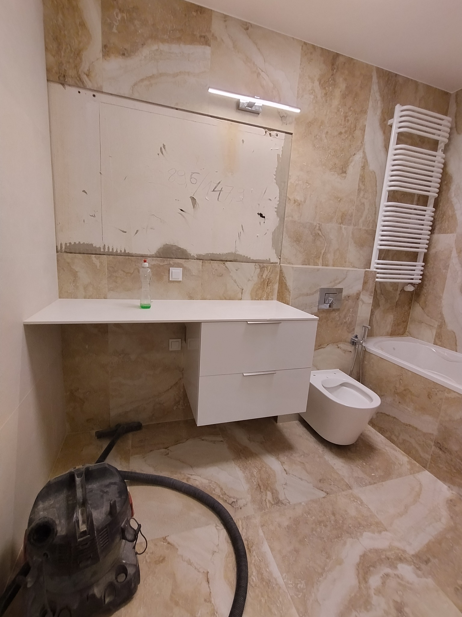 Łazienka w trakcie prac wykończeniowych: częściowo położone marmurowe płytki, biała szafka z umywalką, bidet, drabinka grzejnikowa, odkurzacz przemysłowy na podłodze, ślady po gładzi na ścianie.