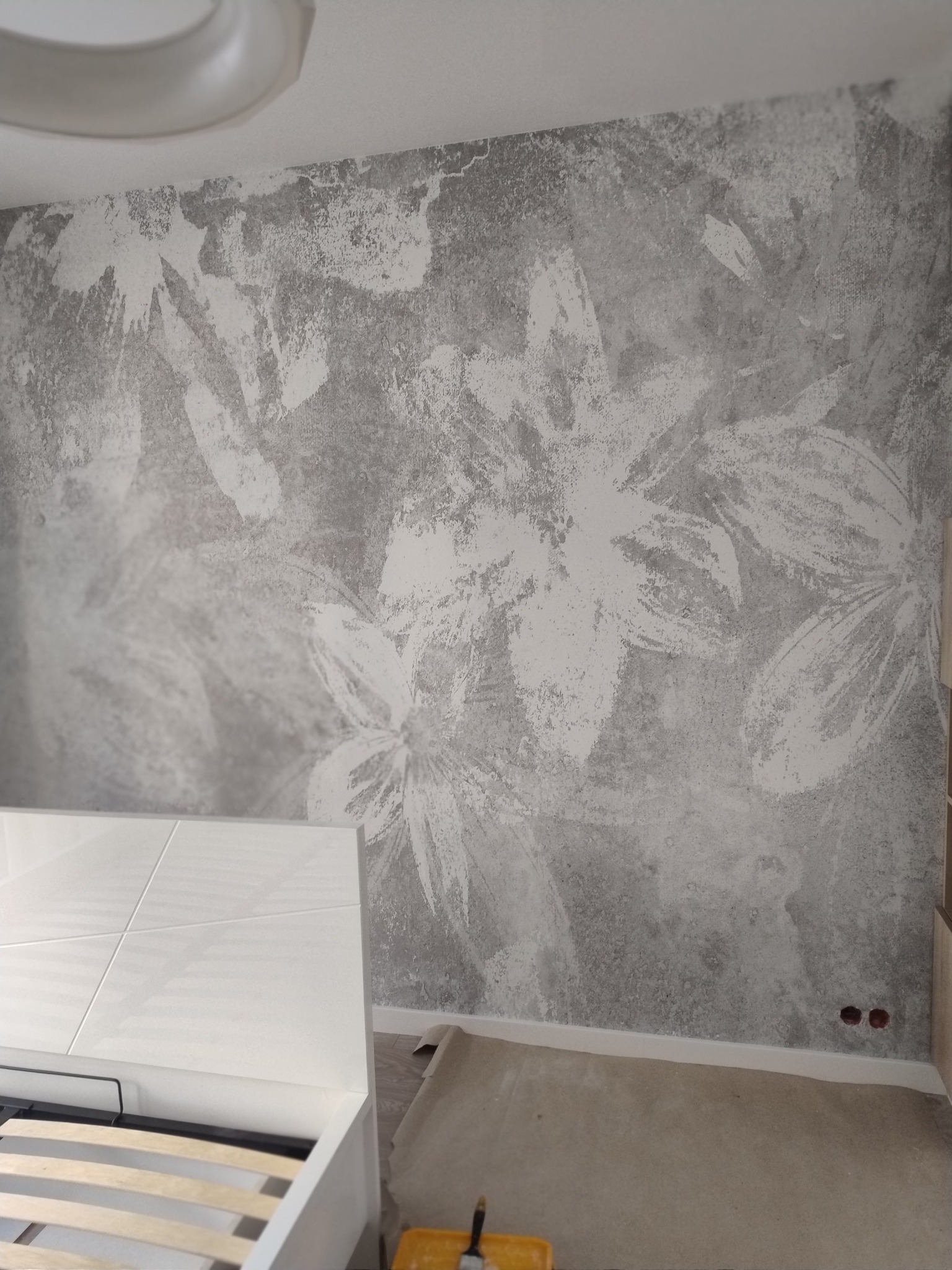 Ściana w pokoju z dekoracyjną szarą gładzią imitującą wzór kwiatowy, widoczny fragment białego łóżka i zabezpieczona podłoga.