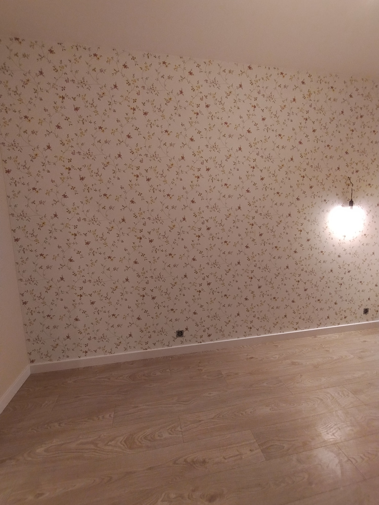 Pokój z tapetą w drobne kwiaty i panelami podłogowymi w odcieniach beżu, widoczna lampa bez abażuru i gniazdko elektryczne.