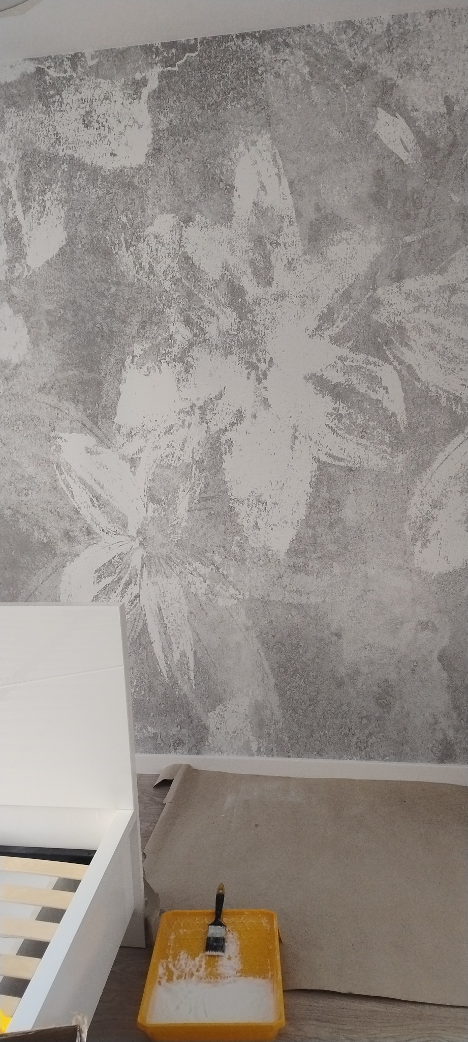 Ściana z dekoracyjną, szarą fakturą imitującą kwiaty, częściowo zasłonięta białym elementem meblowym, z widoczną kuwetą malarską i pędzlem na podłodze zabezpieczonej papierem.