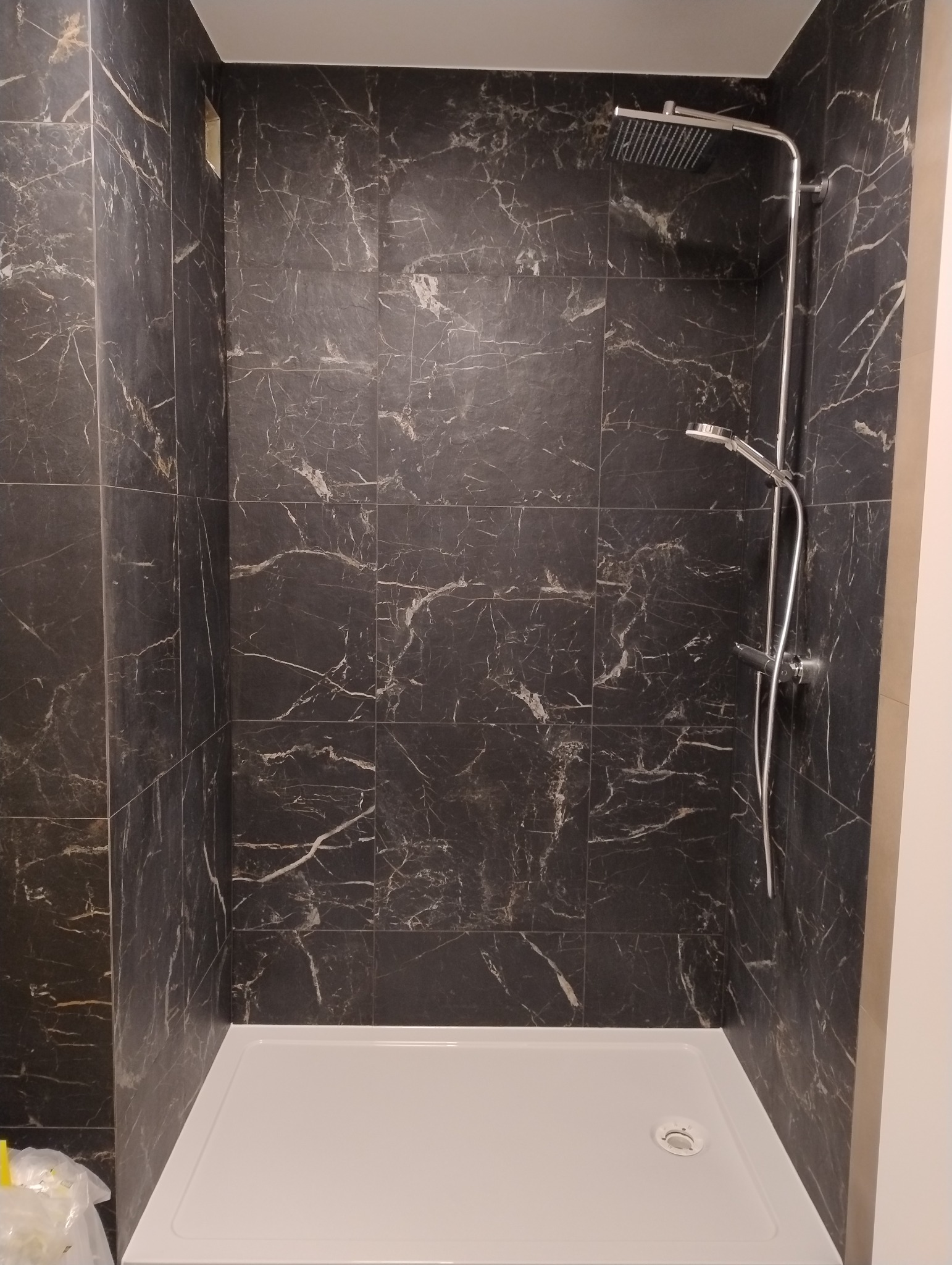 Kabina prysznicowa wyłożona czarnymi płytkami imitującymi marmur z białą brodzikiem i chromowaną armaturą.