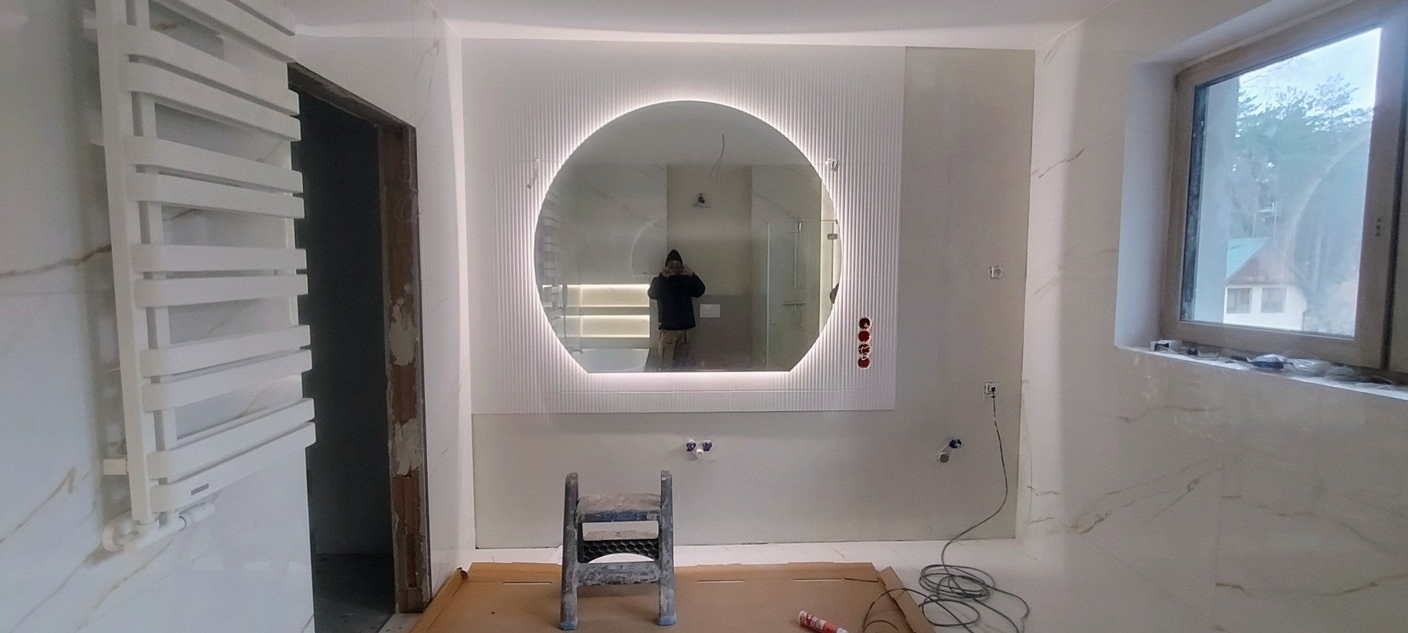 Łazienka w trakcie prac wykończeniowych: białe płytki imitujące marmur, okrągłe lustro z podświetleniem LED, grzejnik drabinkowy, widoczne przewody instalacji elektrycznej i drabina malarska.