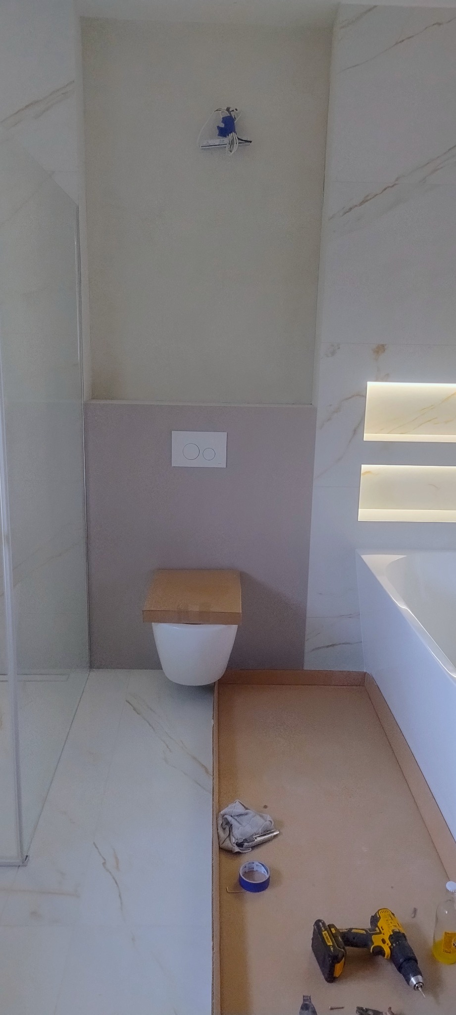 Montaż podwieszanej toalety w łazience z marmurowymi płytkami, widoczne narzędzia na kartonie ochronnym i instalacja elektryczna nad spłuczką.