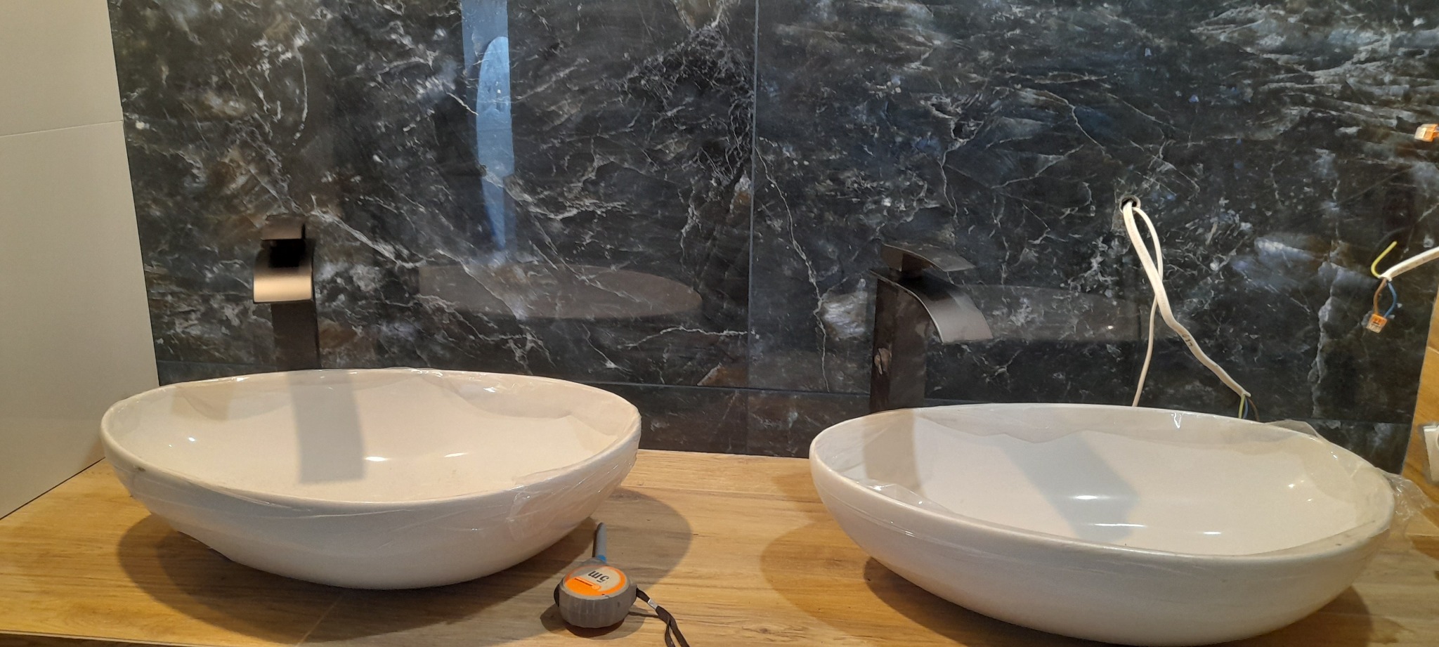 Dwie białe umywalki nablatowe, jeszcze w folii ochronnej, stoją na drewnianym blacie. W tle ściana wyłożona czarnymi płytkami imitującymi marmur z zamontowanymi bateriami. Widoczne przewody...
