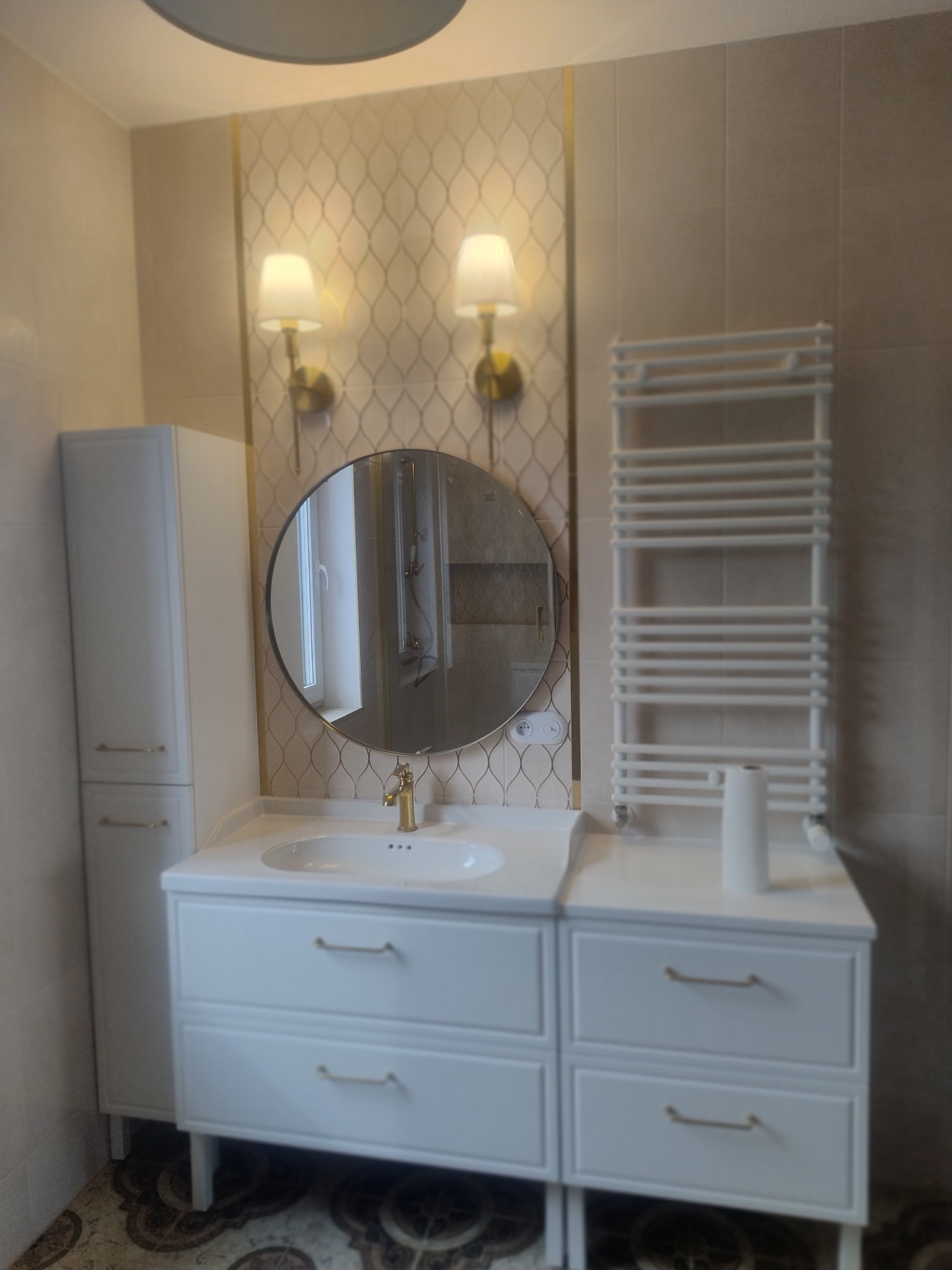 Wykończona łazienka z białą szafką pod umywalkę z złotymi uchwytami, okrągłym lustrem z widocznym odbiciem kabiny prysznicowej, grzejnikiem drabinkowym i dekoracyjną tapetą w odcieniach beżu i złota.