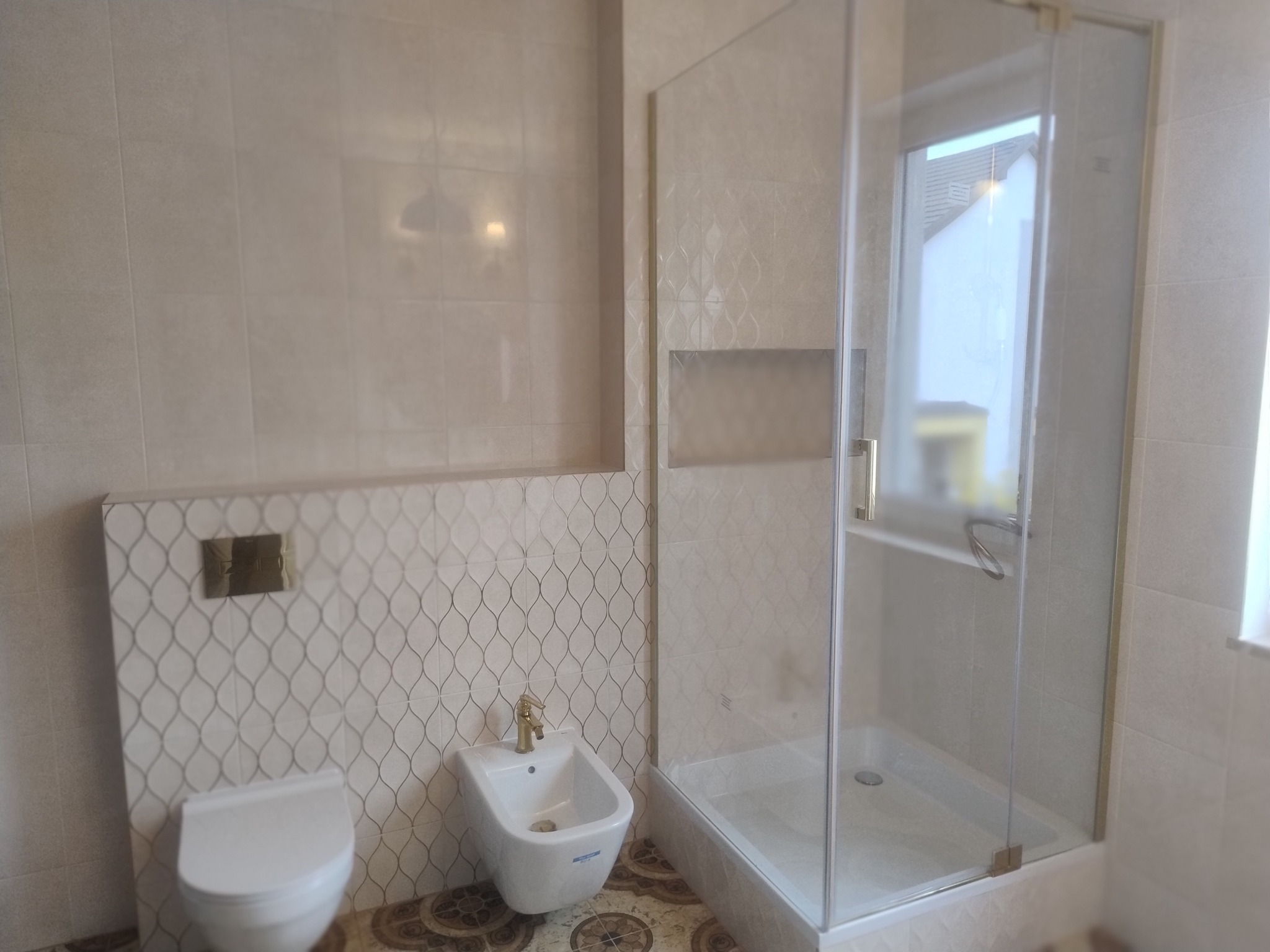 Jasna łazienka z kabiną prysznicową z przezroczystego szkła, bidetem z baterią w kolorze złota, toaletą podwieszaną i płytkami ściennymi w odcieniach beżu z delikatnym wzorem.