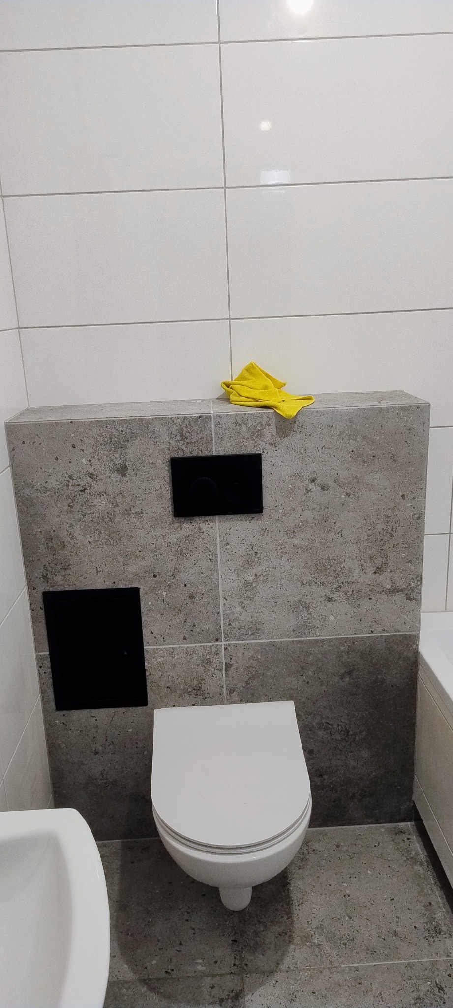 Nowoczesna toaleta podwieszana z szarą obudową, czarnymi przyciskami spłukiwania i żółtą ściereczką na górze, na tle białych płytek