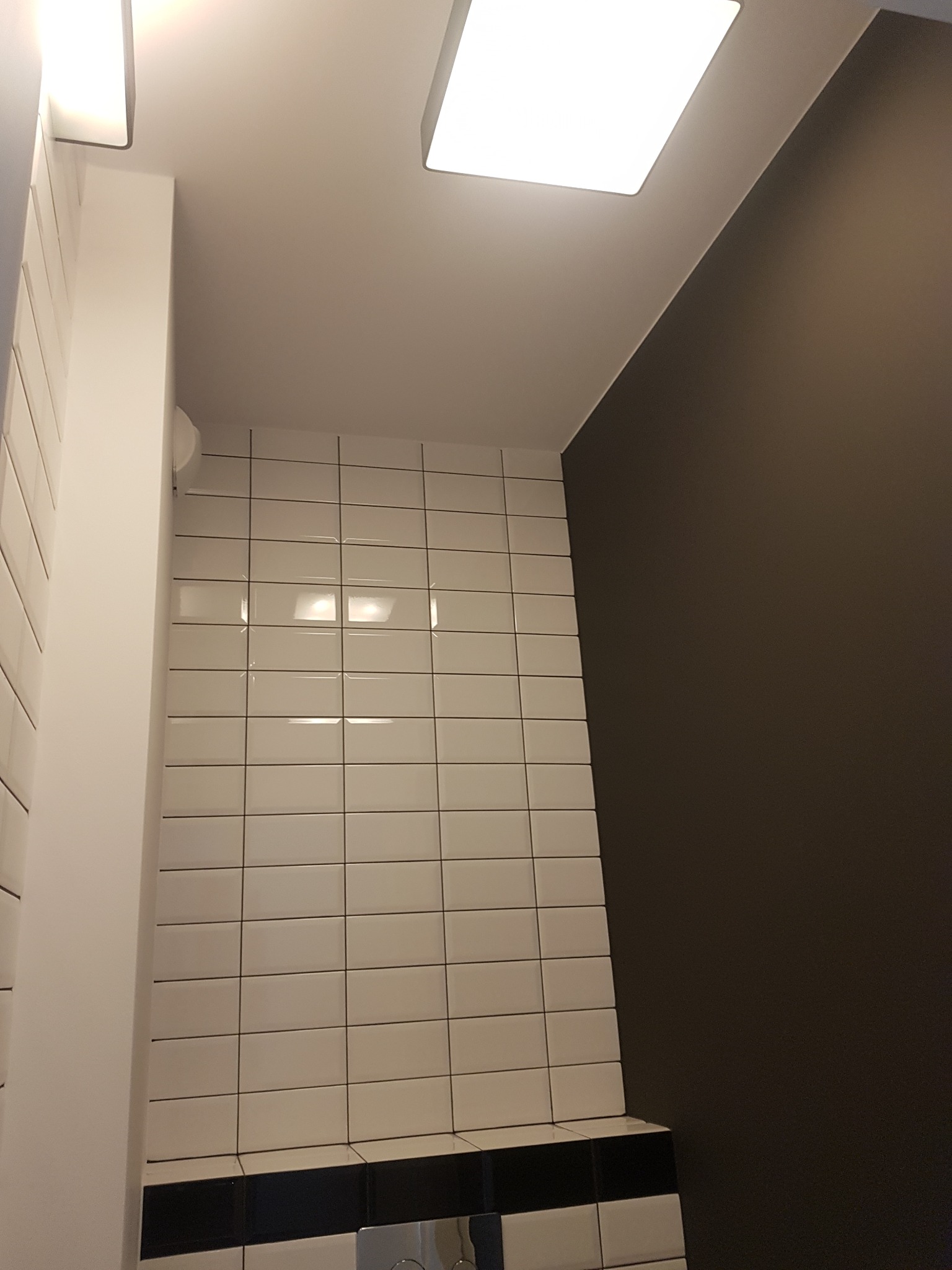 Białe, prostokątne płytki na ścianie łazienki z ciemną fugą, kontrastujące z ciemnoszarą ścianą obok, oświetlone kwadratową lampą sufitową.