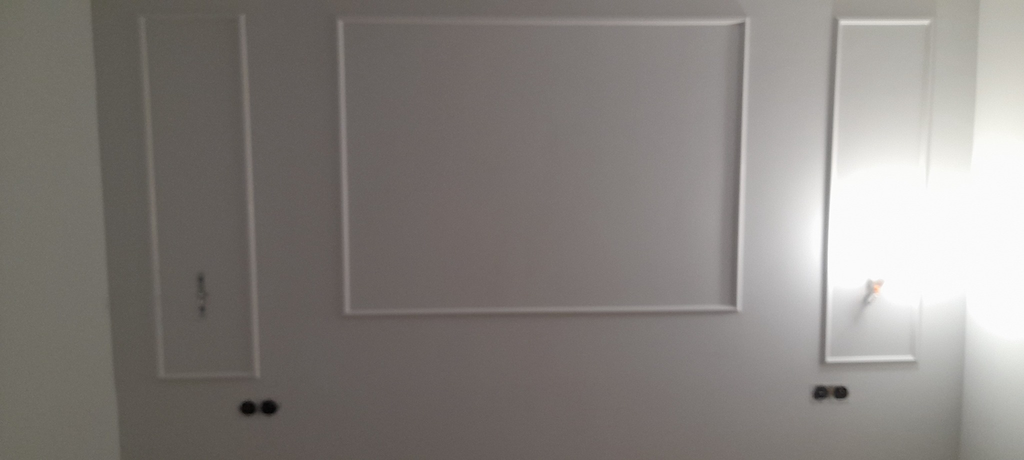 Szara ściana z dekoracyjnymi, prostokątnymi ramkami i gniazdkami elektrycznymi w trakcie montażu.