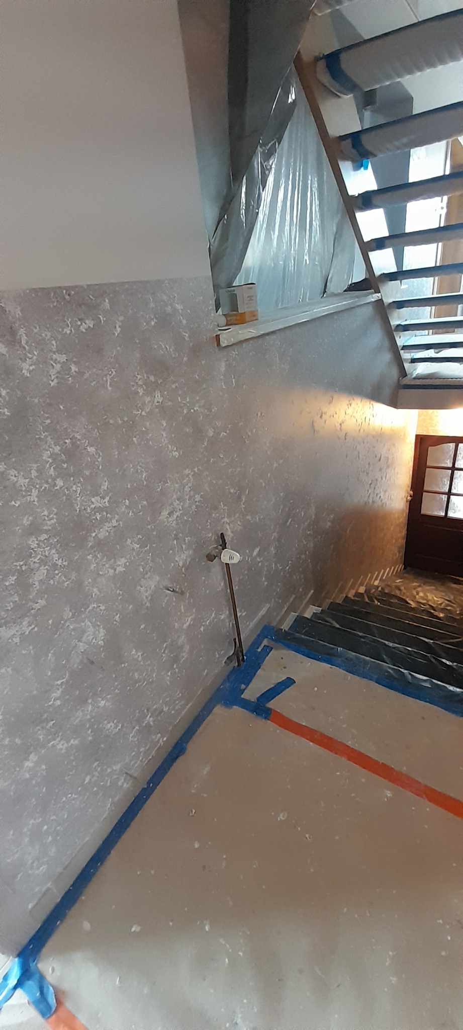 Nakładanie gładzi dekoracyjnej na ścianie klatki schodowej, zabezpieczone schody i podłoga, widoczny pojemnik z farbą i metalowa rurka przy ścianie.