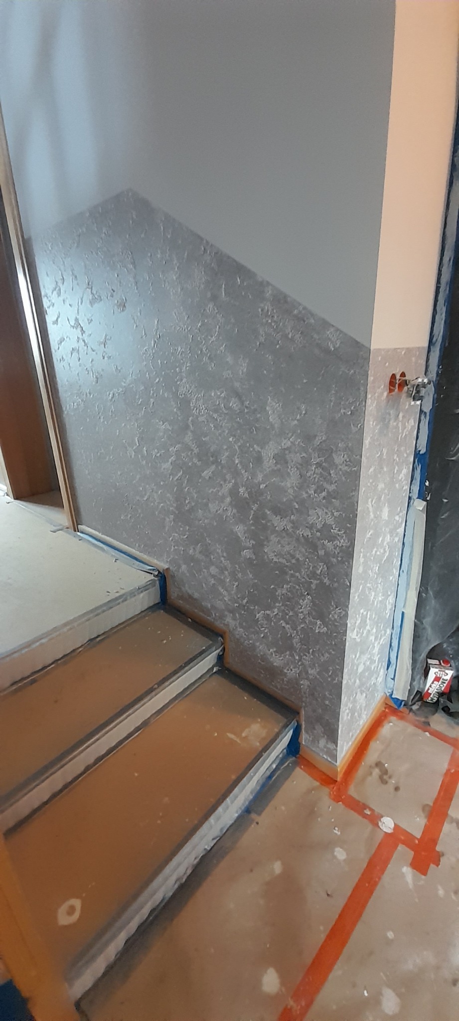 Ściana z dekoracyjną szarą fakturą przy schodach zabezpieczonych folią malarską, widoczne gniazdka elektryczne i taśmy malarskie.