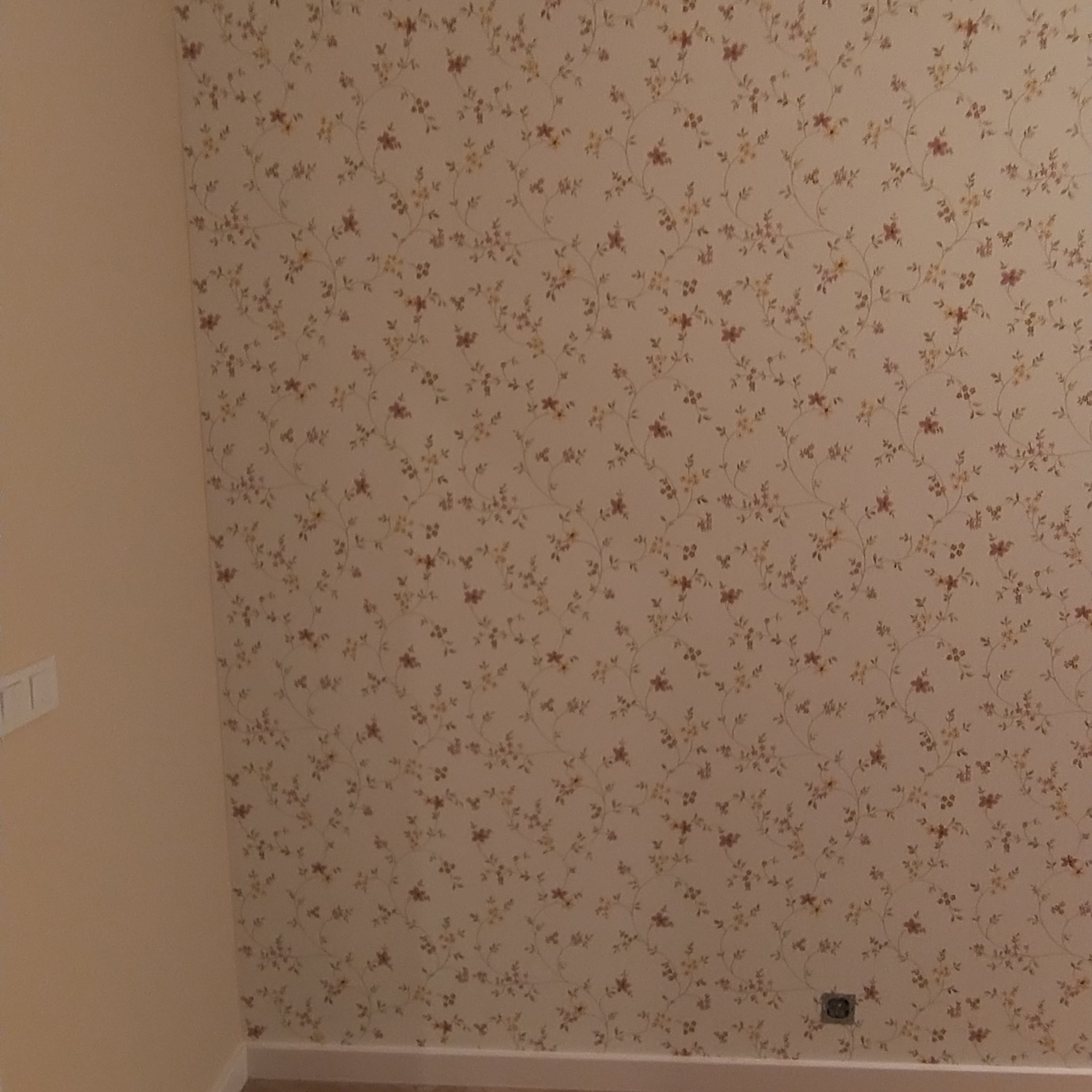 Ściana pokryta tapetą w drobne kwiatowe wzory w odcieniach beżu, brązu i żółci, widoczna obok gładkiej, jasnej ściany z włącznikiem światła.