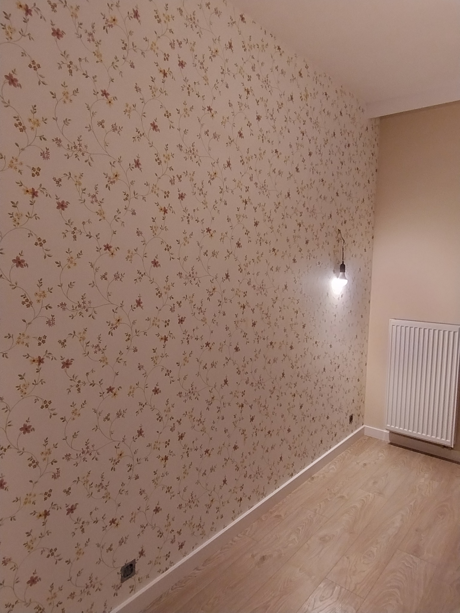 Ściana z tapetą w drobne kwiaty, widoczna instalacja elektryczna z żarówką i grzejnik na ścianie w wykończonym pokoju.