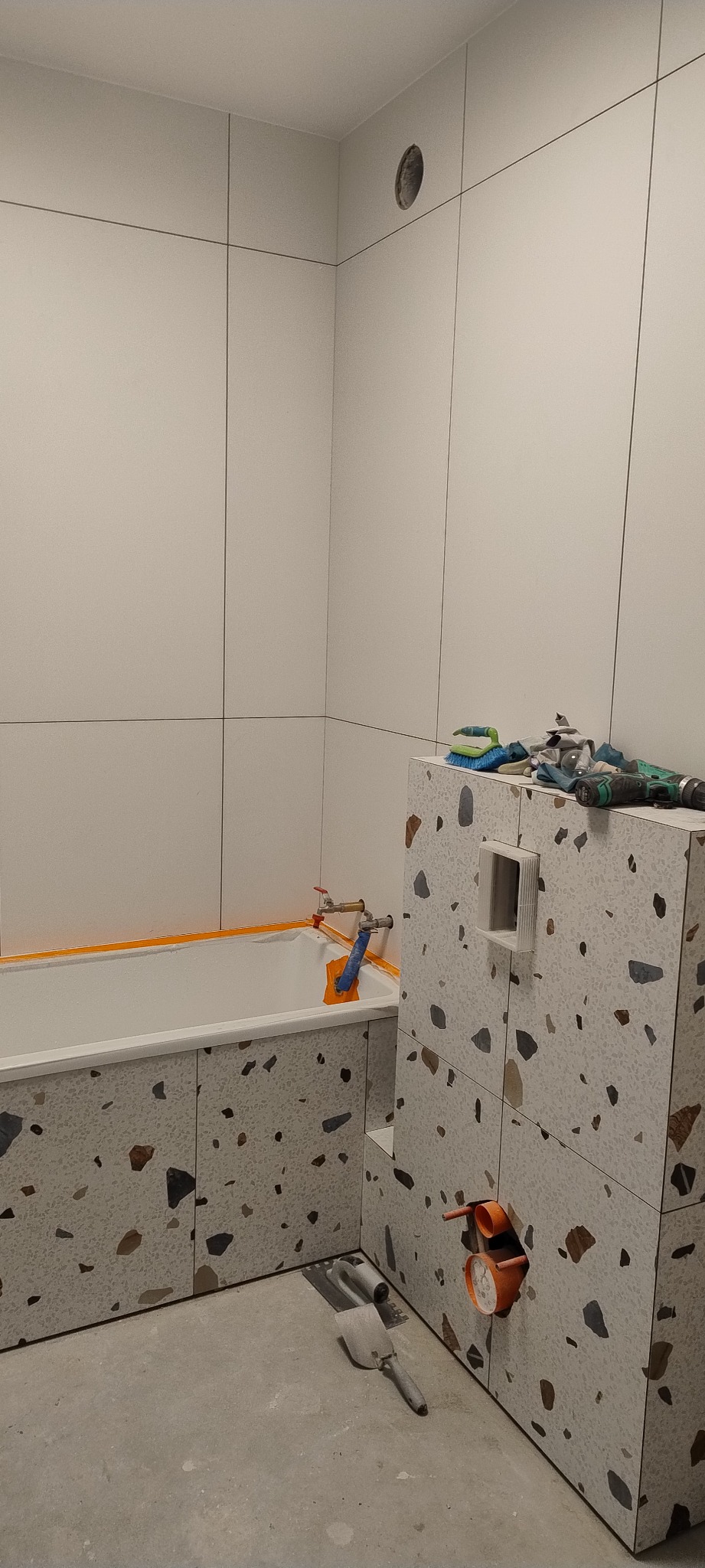 Łazienka w trakcie wykańczania: wanna obudowana płytkami lastryko, białe płytki na ścianach, widoczne narzędzia glazurnicze i otwór na wentylację.