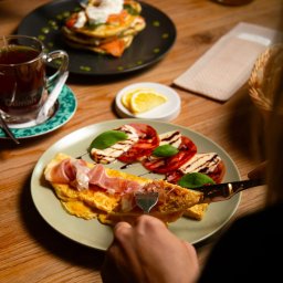 GRANA FOOD REST 1 SPÓŁKA Z OGRANICZONĄ ODPOWIEDZIALNOŚCIĄ - Osoba kroi omlet z szynką parmeńską na talerzu, obok sałatka caprese i filiżanka herbaty Dilmah, w tle pancakes z łososiem i jajkiem w koszulce.