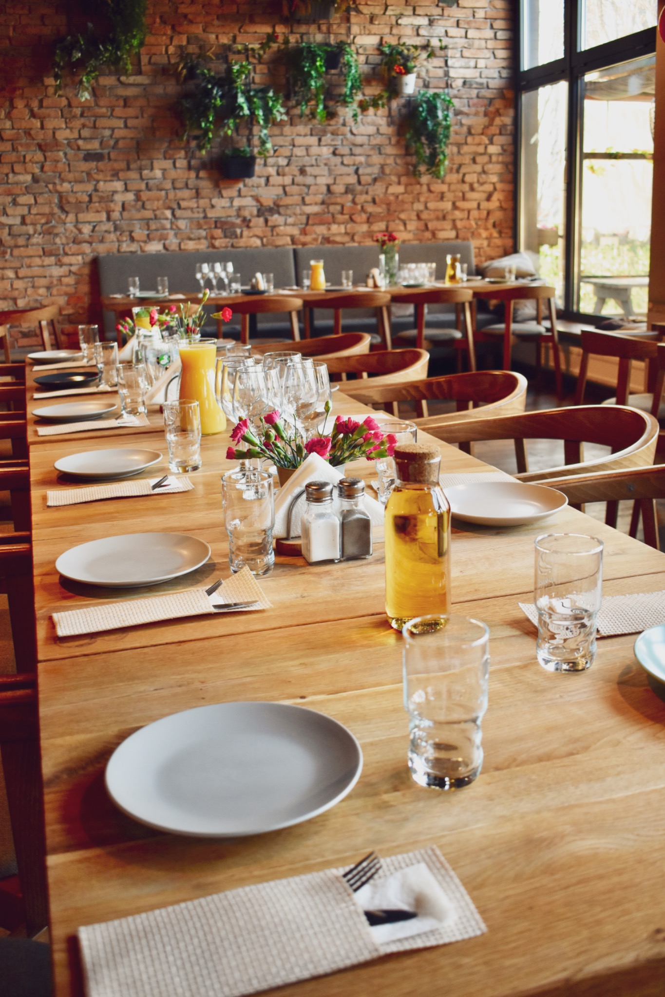 Długi stół z drewna, nakryty szarymi talerzami, szklankami, sztućcami w serwetkach, butelkami z oliwą i małymi bukietami różowych kwiatów, ustawiony w restauracji z ceglaną ścianą i zielonymi...
