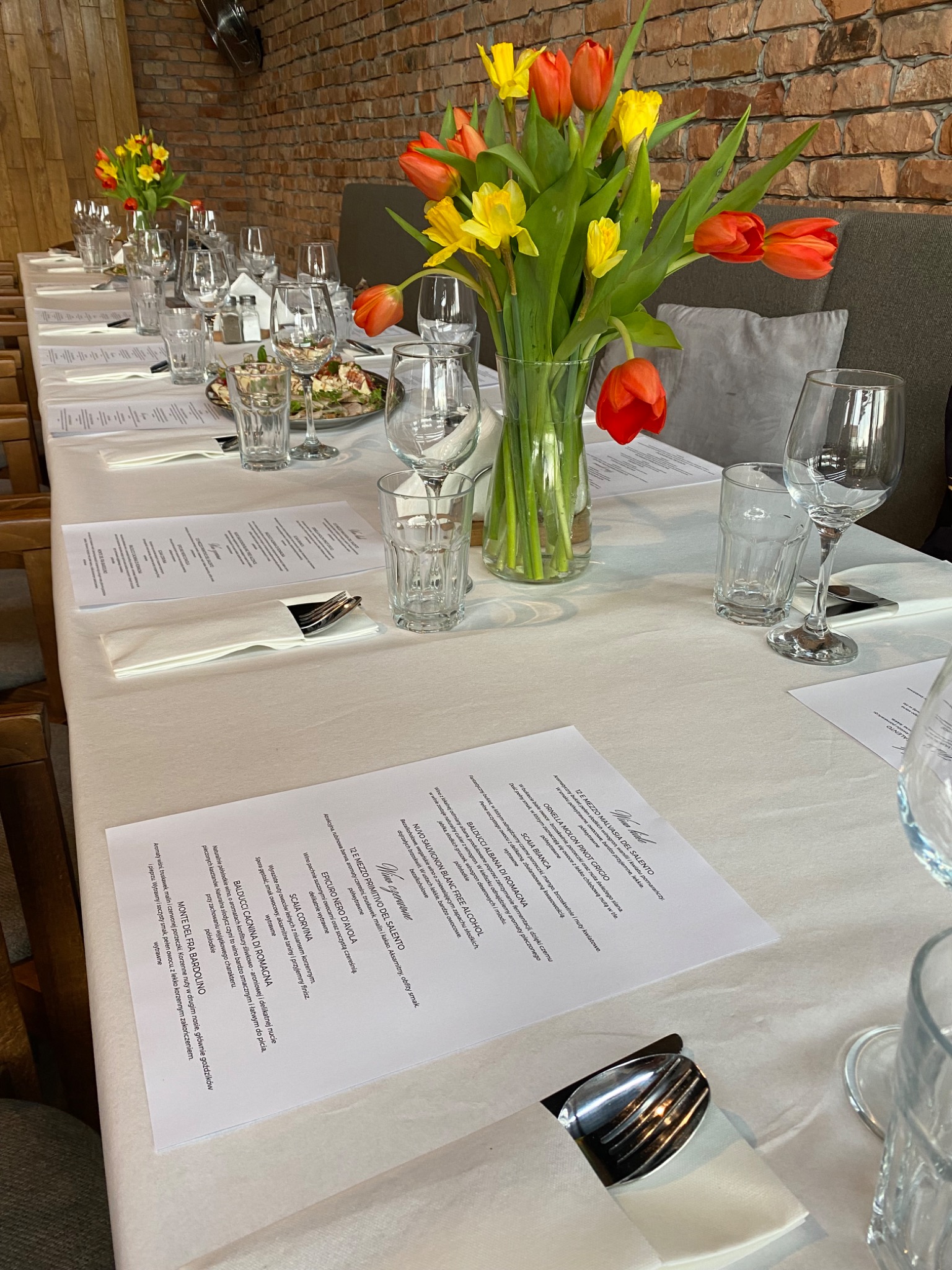 Uroczysty stół z białym obrusem, nakryty zastawą, kieliszkami, sztućcami i menu, ozdobiony wazonem z żółto-pomarańczowymi tulipanami na tle ceglanej ściany.