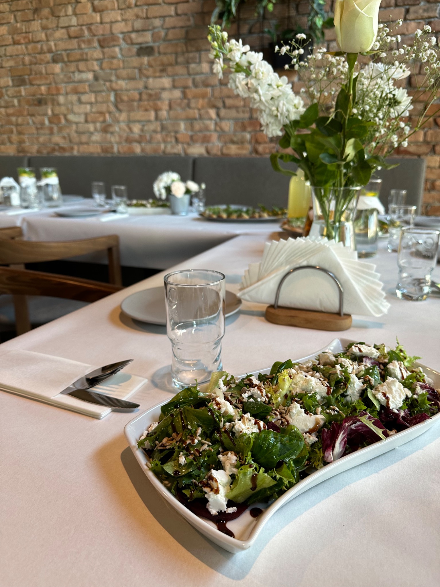 Stół z białym obrusem, na którym znajduje się sałatka z serem i sosem balsamicznym w białym naczyniu, sztućce, szklanka oraz dekoracje kwiatowe w wazonie, w tle ceglana ściana i nakryte stoły...