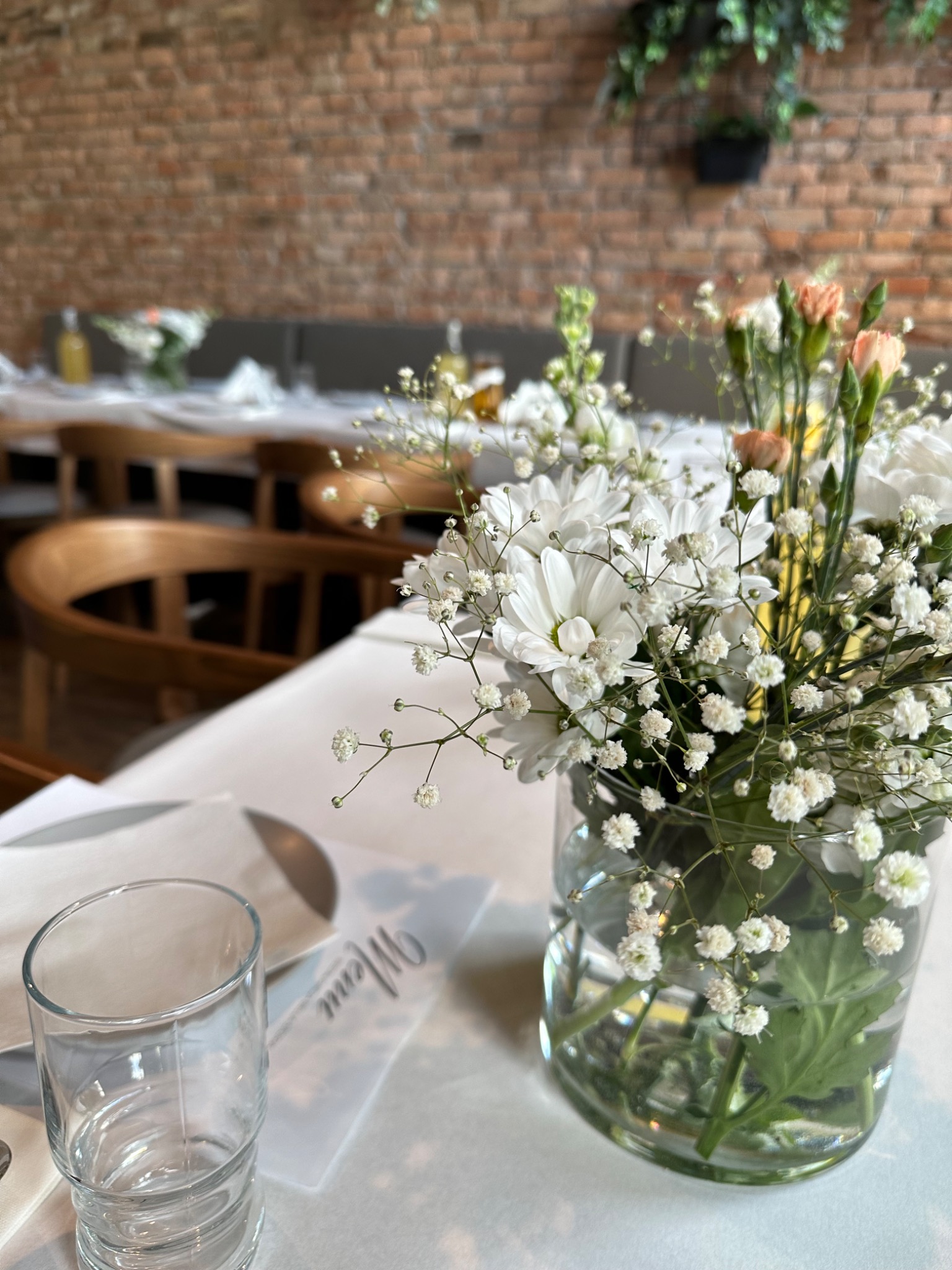 Dekoracja stołu z białych kwiatów i gipsówki w szklanym wazonie, ustawiona na białym obrusie obok pustej szklanki i karty menu, w tle ceglana ściana i nakryte stoły.