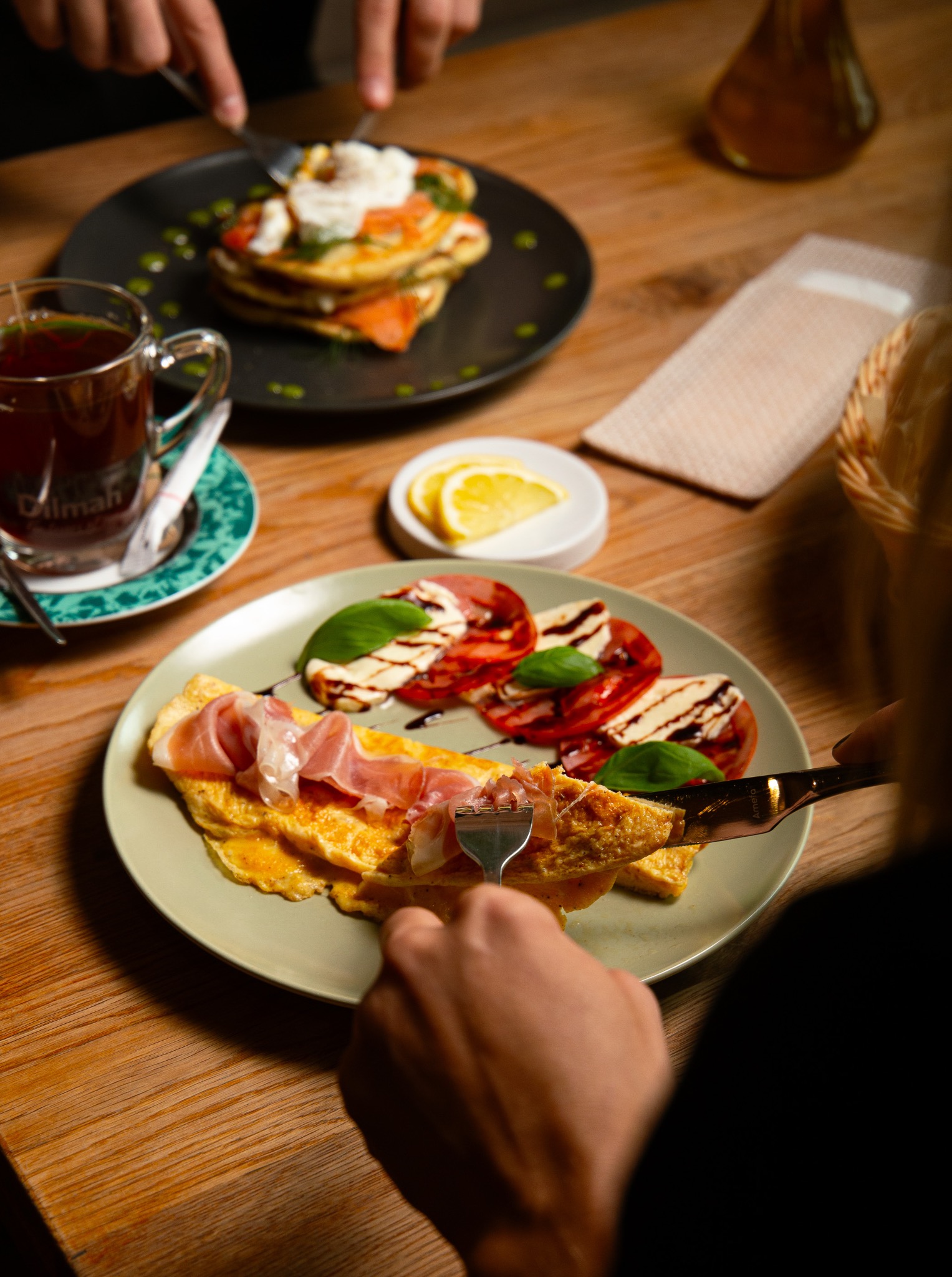 Osoba kroi omlet z szynką parmeńską na talerzu, obok sałatka caprese i filiżanka herbaty Dilmah, w tle pancakes z łososiem i jajkiem w koszulce.