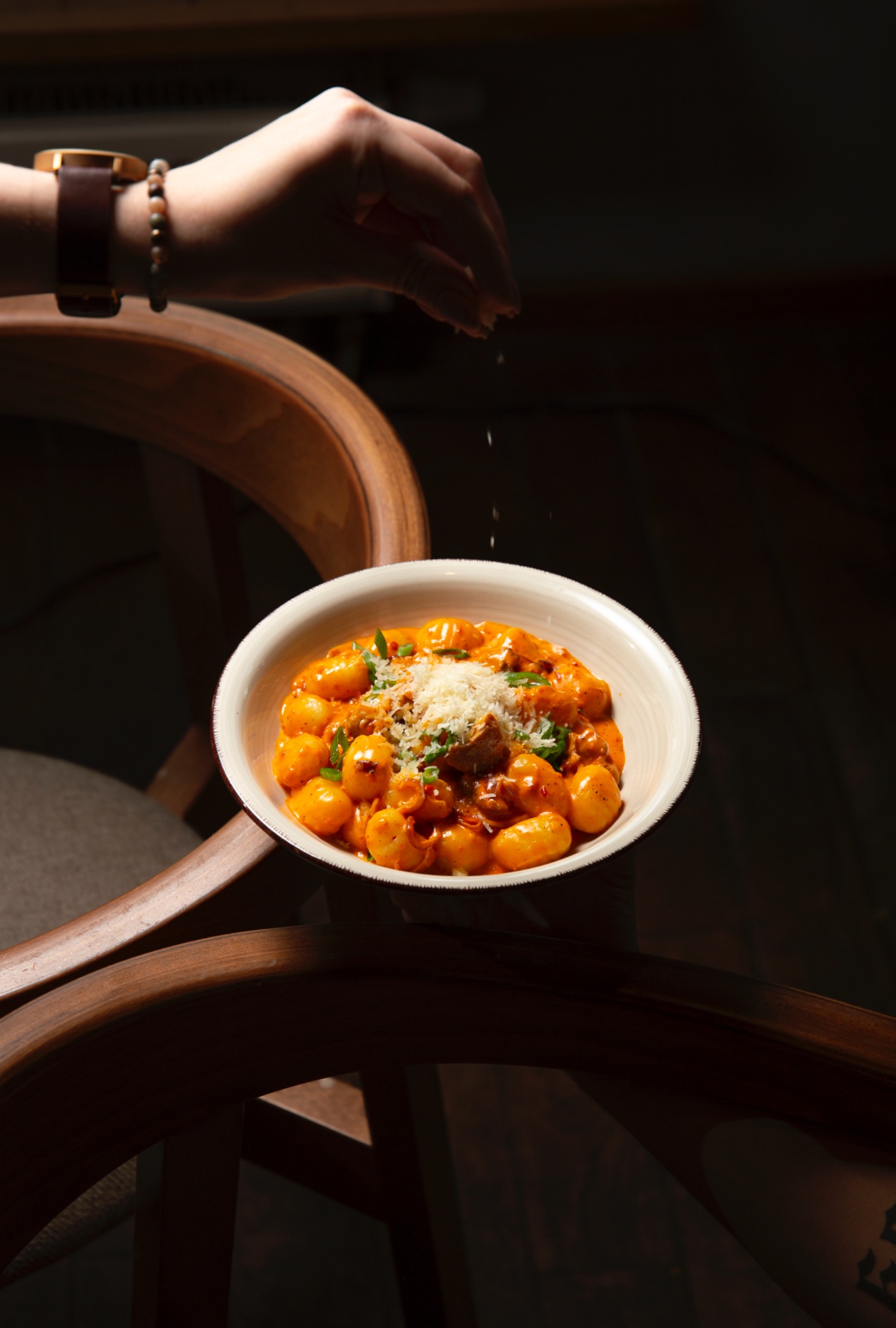 Gnocchi w pomarańczowym sosie, posypywane serem z dłoni, podane na drewnianym krześle w restauracji.