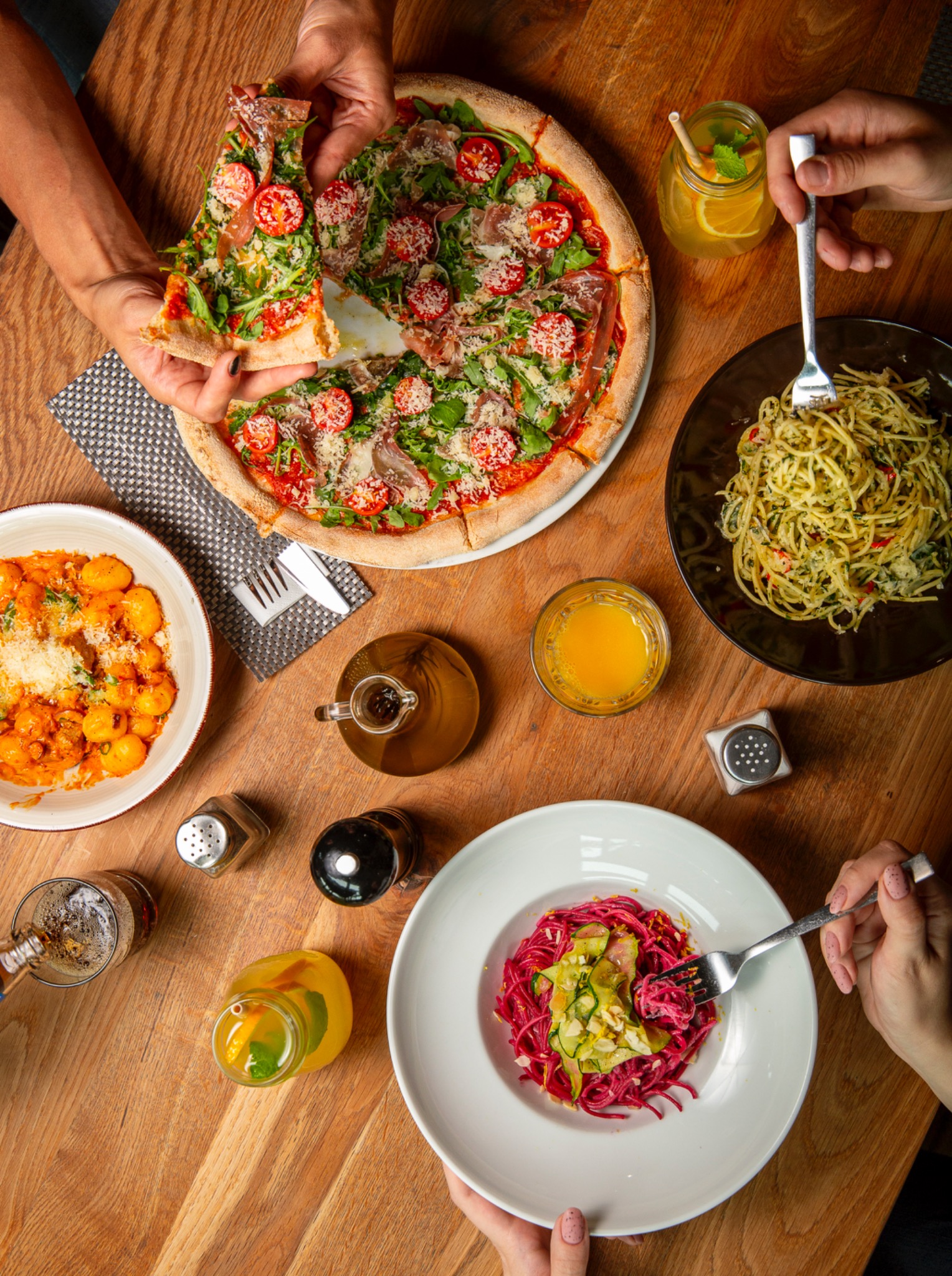 Widok z góry na stół z pizzą z rukolą i prosciutto, gnocchi w sosie pomidorowym, zielone spaghetti i czerwone spaghetti z cukinią, uzupełniony napojami w szklankach i butelkach, z widocznymi dłońmi...