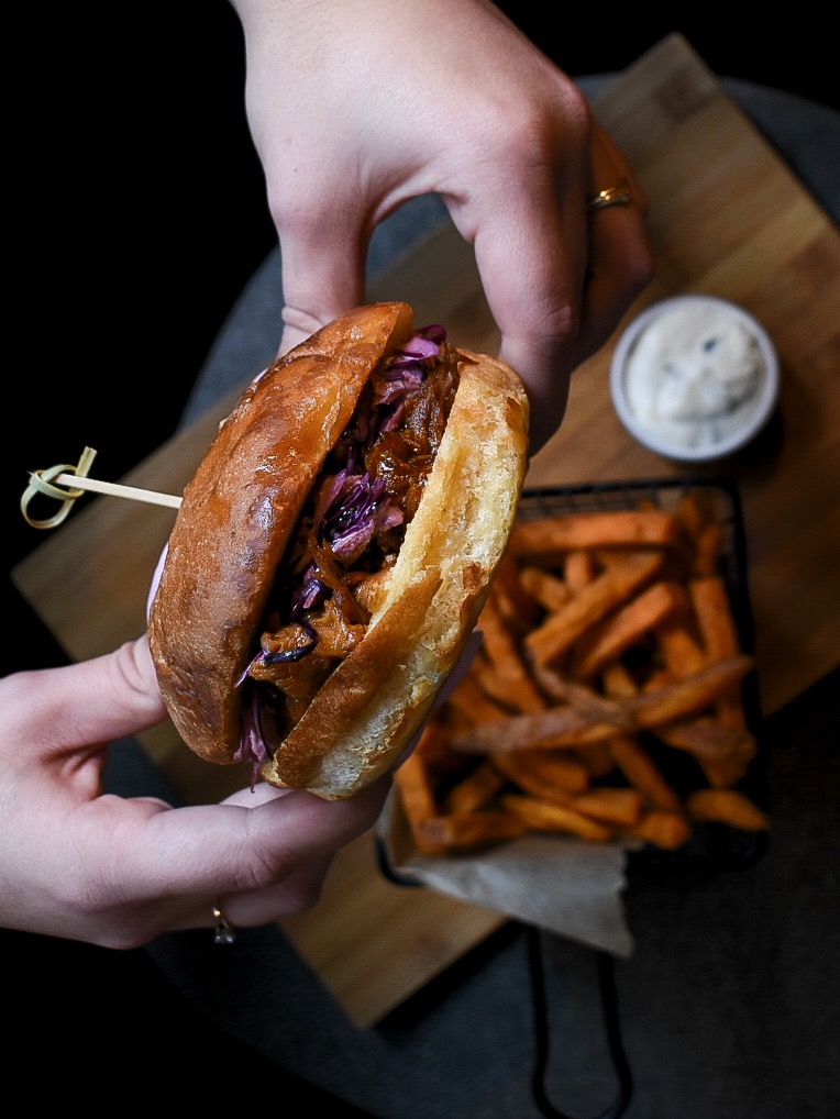 Pulled jackfruit burger z czerwoną kapustą, podawany z frytkami z batatów i sosem, trzymany w dłoniach z pierścionkami na palcach, na drewnianej desce.