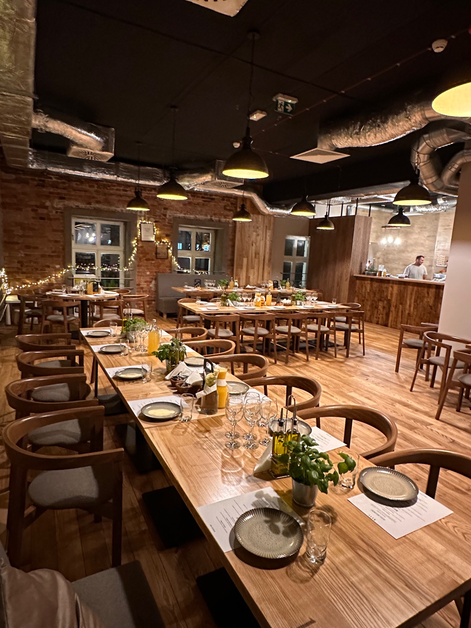 Sala restauracyjna z drewnianymi stołami i krzesłami, nakryta zastawa, oświetlenie punktowe, ceglana ściana i widoczny fragment baru z obsługą.