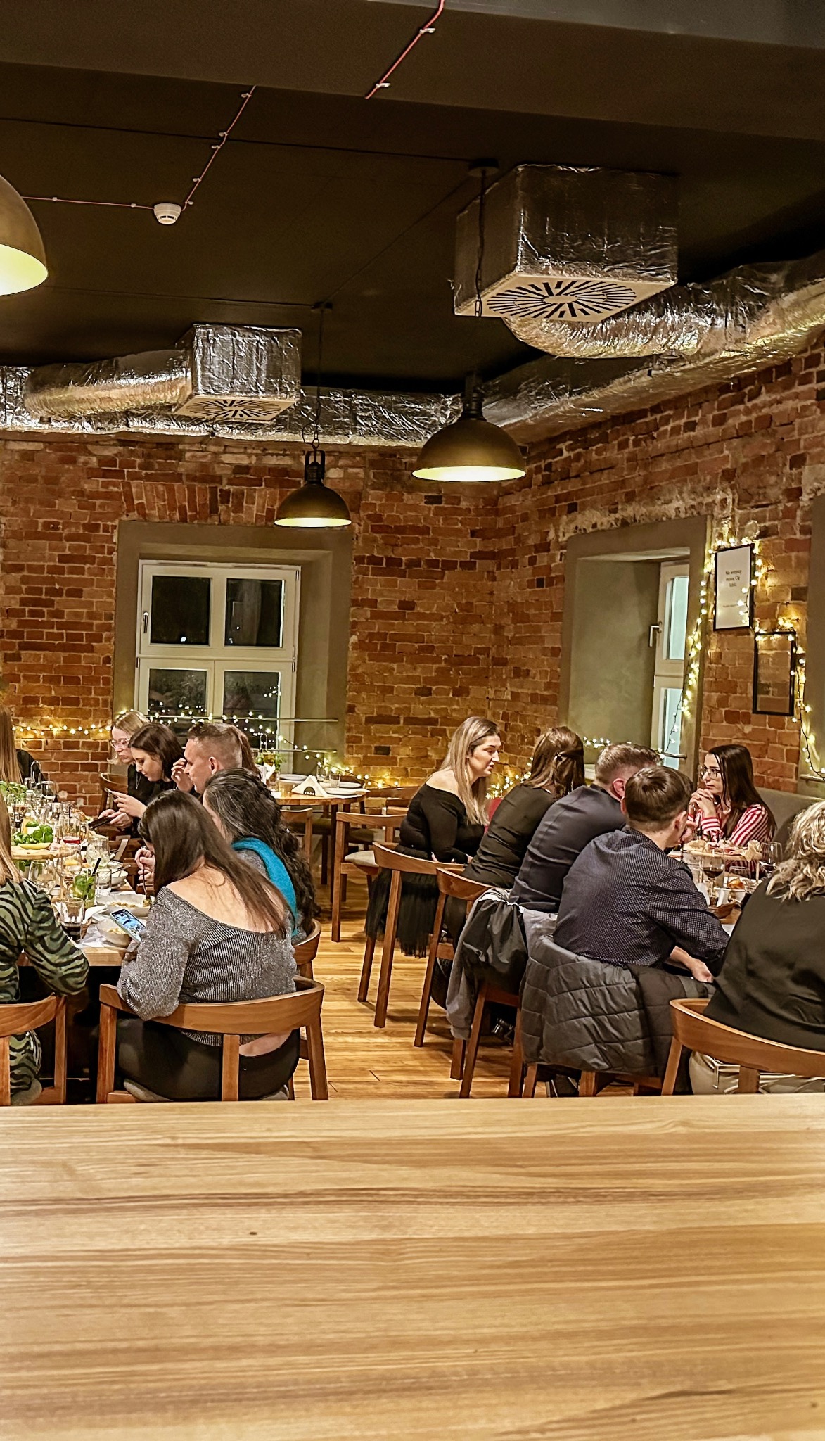 Uczestnicy siedzą przy drewnianych stołach podczas wydarzenia w ceglanej sali z dekoracyjnym oświetleniem i widocznymi elementami wentylacji na suficie.