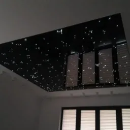 Czarny, podwieszany sufit z efektem rozgwieżdżonego nieba w pomieszczeniu z oknem i roletami.