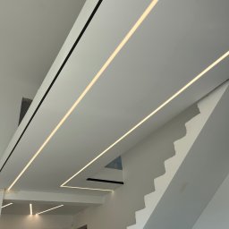 Artur Kalinin sufity napinanie PROSPECT - Nowoczesny biały sufit z wbudowanym oświetleniem liniowym LED i czarnymi akcentami, tworzący geometryczny wzór, widoczny w Alwerni. Częściowo widoczne schody.