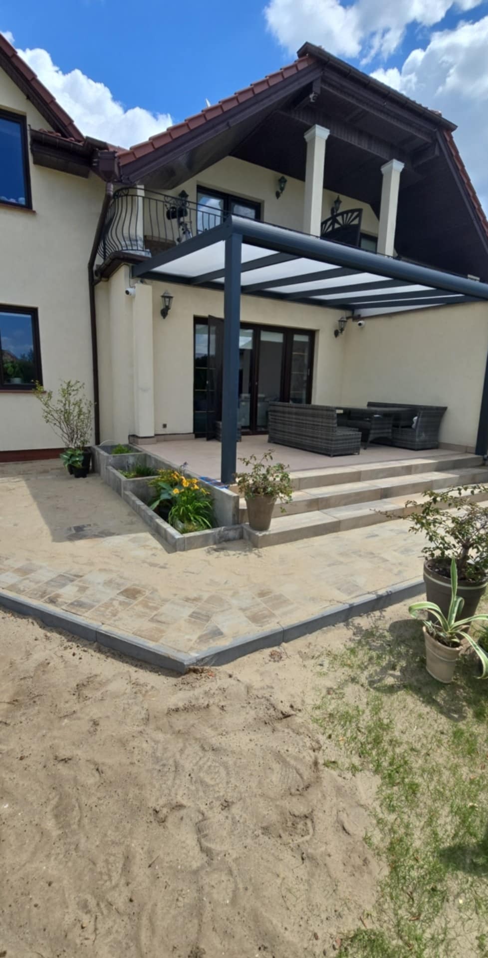 Wykończony taras z kostki brukowej z podwyższonymi rabatami kwiatowymi i pergolą, idealny na letni wypoczynek w ogrodzie.