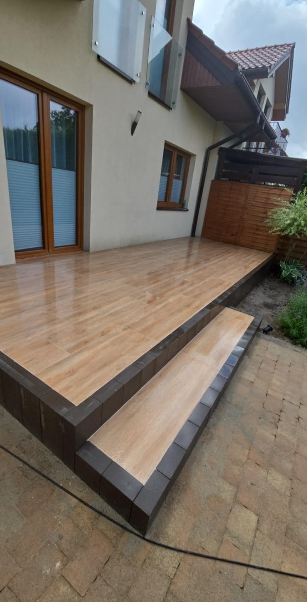 Wykończony taras z płytek imitujących drewno, z dwoma schodkami, obrzeża z ciemnej kostki. Widok na elewację domu z drewnianą stolarką okienną i szklanymi balustradami.