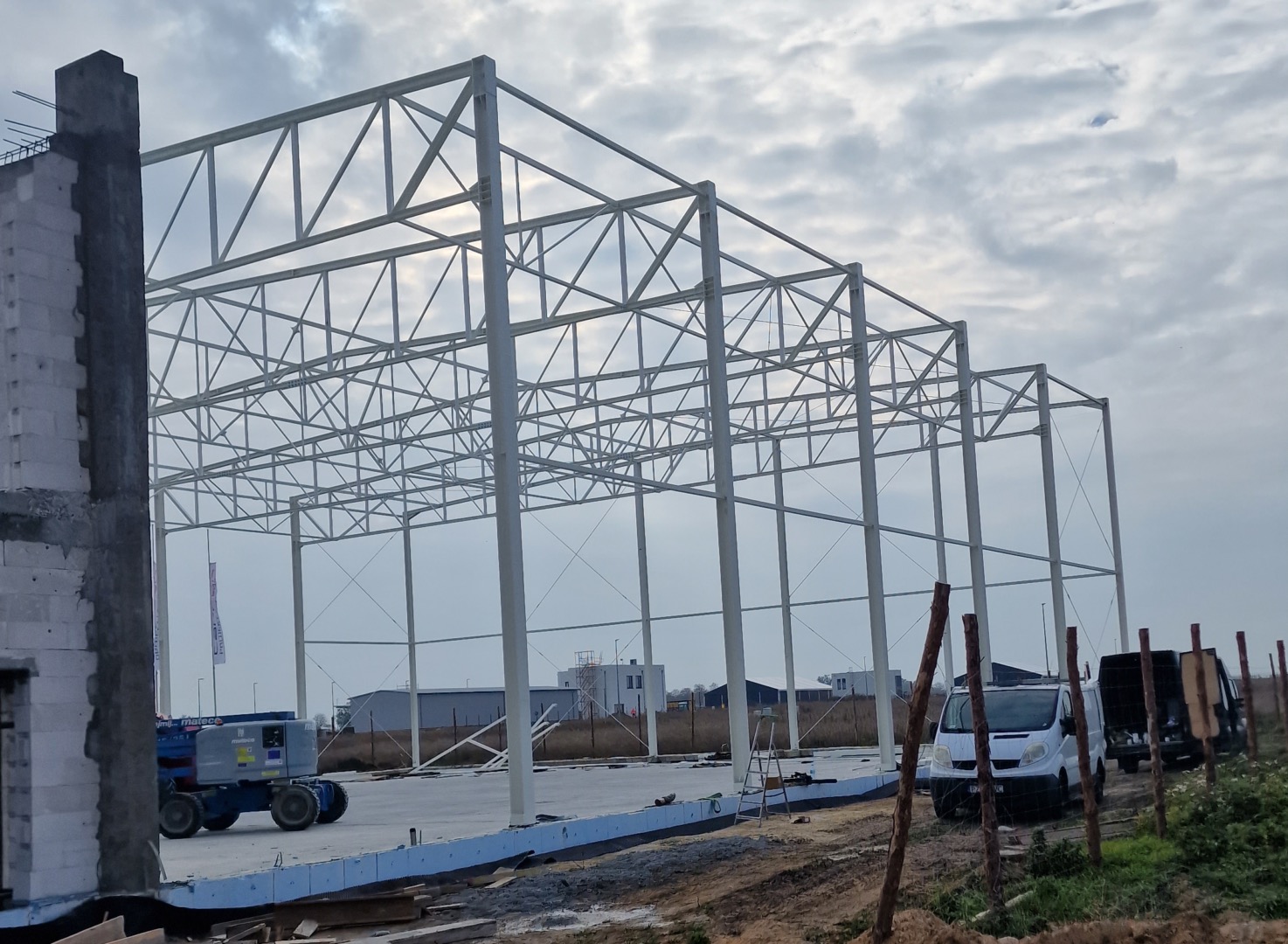 Stalowa konstrukcja hali w trakcie budowy, widoczna z zewnątrz, z podnośnikiem koszowym i samochodami dostawczymi w tle, betonowa podstawa w trakcie wylewania.