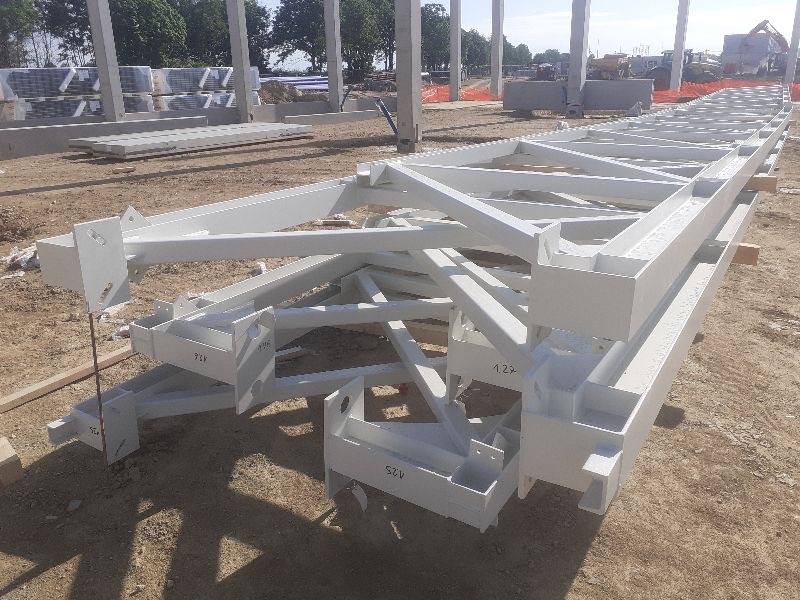 Biała, stalowa konstrukcja kratowa leżąca na ziemi placu budowy, z widocznymi betonowymi słupami w tle i oznaczeniami numeracyjnymi na elementach.