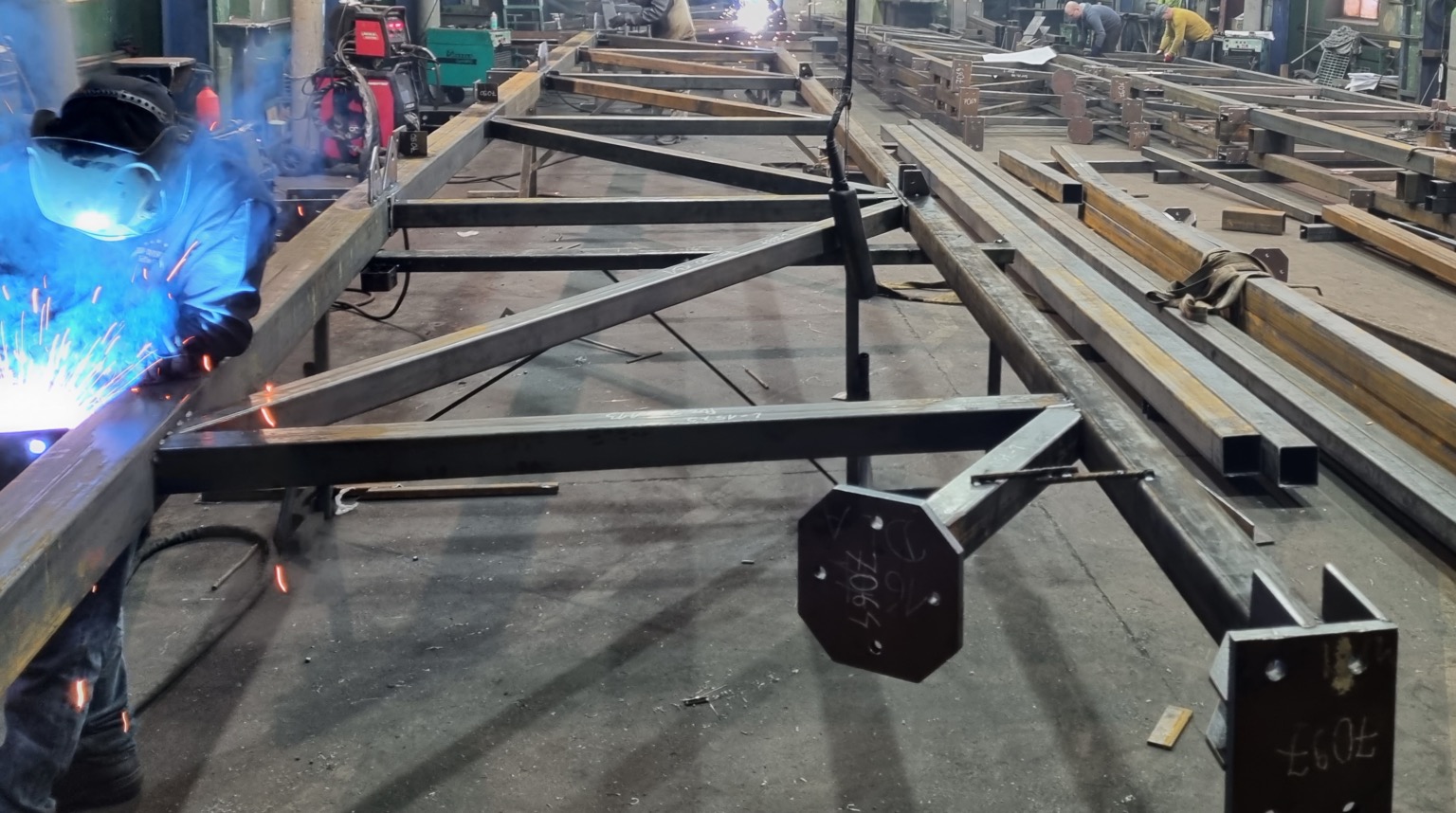 Pracownik w kombinezonie spawalniczym łączy elementy konstrukcji stalowej za pomocą spawarki, widoczne iskry i niebieskie światło łuku elektrycznego, w tle hala produkcyjna z ułożonymi elementami...