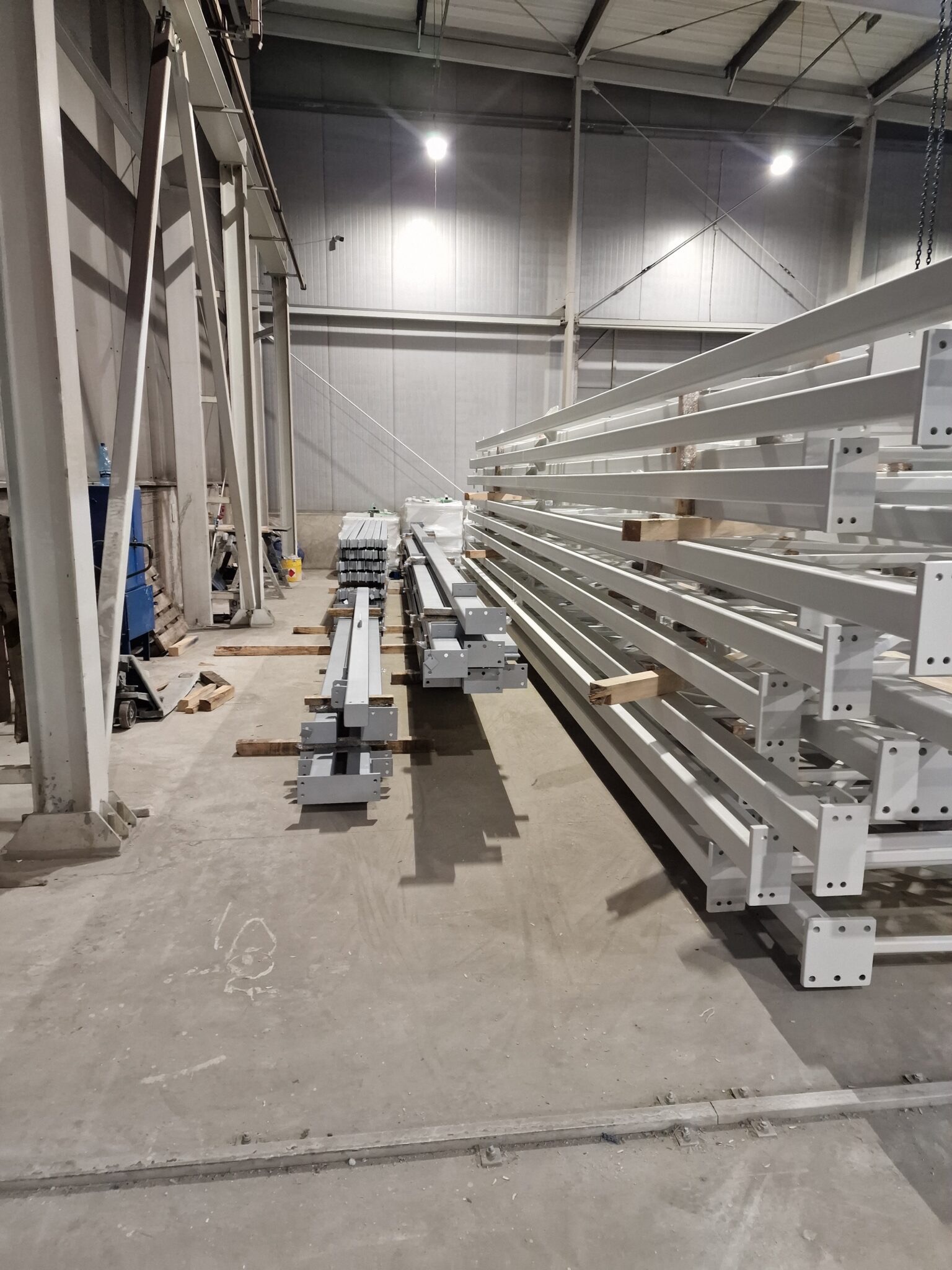 Stalowe, pomalowane na biało elementy konstrukcyjne hali magazynowane wewnątrz budynku, widoczne ślady po wózku widłowym na betonowej posadzce.