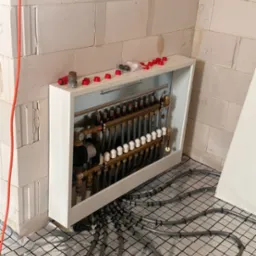 Rozdzielacz ogrzewania podłogowego z widocznymi rurami rozprowadzonymi na folii izolacyjnej, w tle ściana z betonu kom&oacute;rkowego.