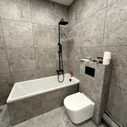 Hydraulika Maciej Michałkiewicz - Wykończona łazienka w Środzie Wielkopolskiej z wanną, toaletą podwieszaną i deszczownicą, wyłożona szarymi płytkami imitującymi beton, z widocznymi rurami kanalizacyjnymi.