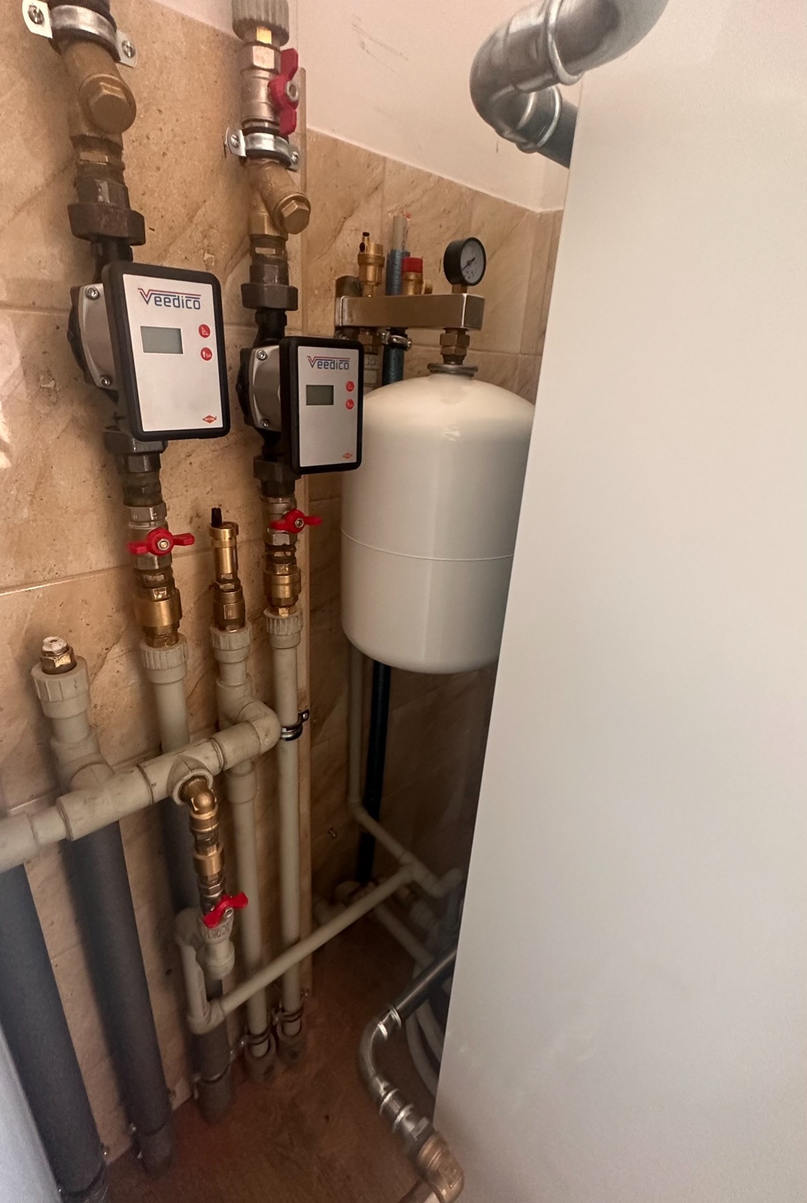 Instalacja grzewcza z dwoma elektronicznymi pompami obiegowymi Veedico, zbiornikiem wyrównawczym i rurami miedzianymi oraz z tworzyw sztucznych na tle beżowej ściany z płytek.