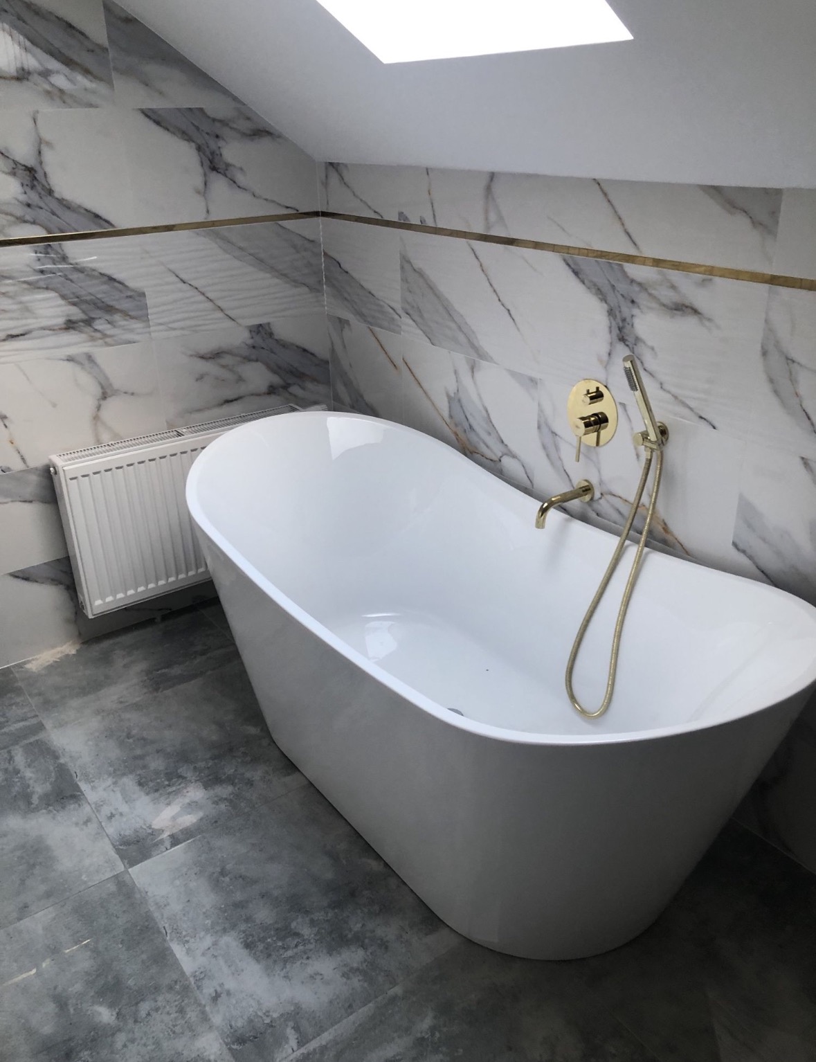 Wolnostojąca wanna z baterią w kolorze złotym na tle marmurowych płytek z dekoracyjną listwą, grzejnik żeberkowy z boku, podłoga wyłożona szarymi płytkami.